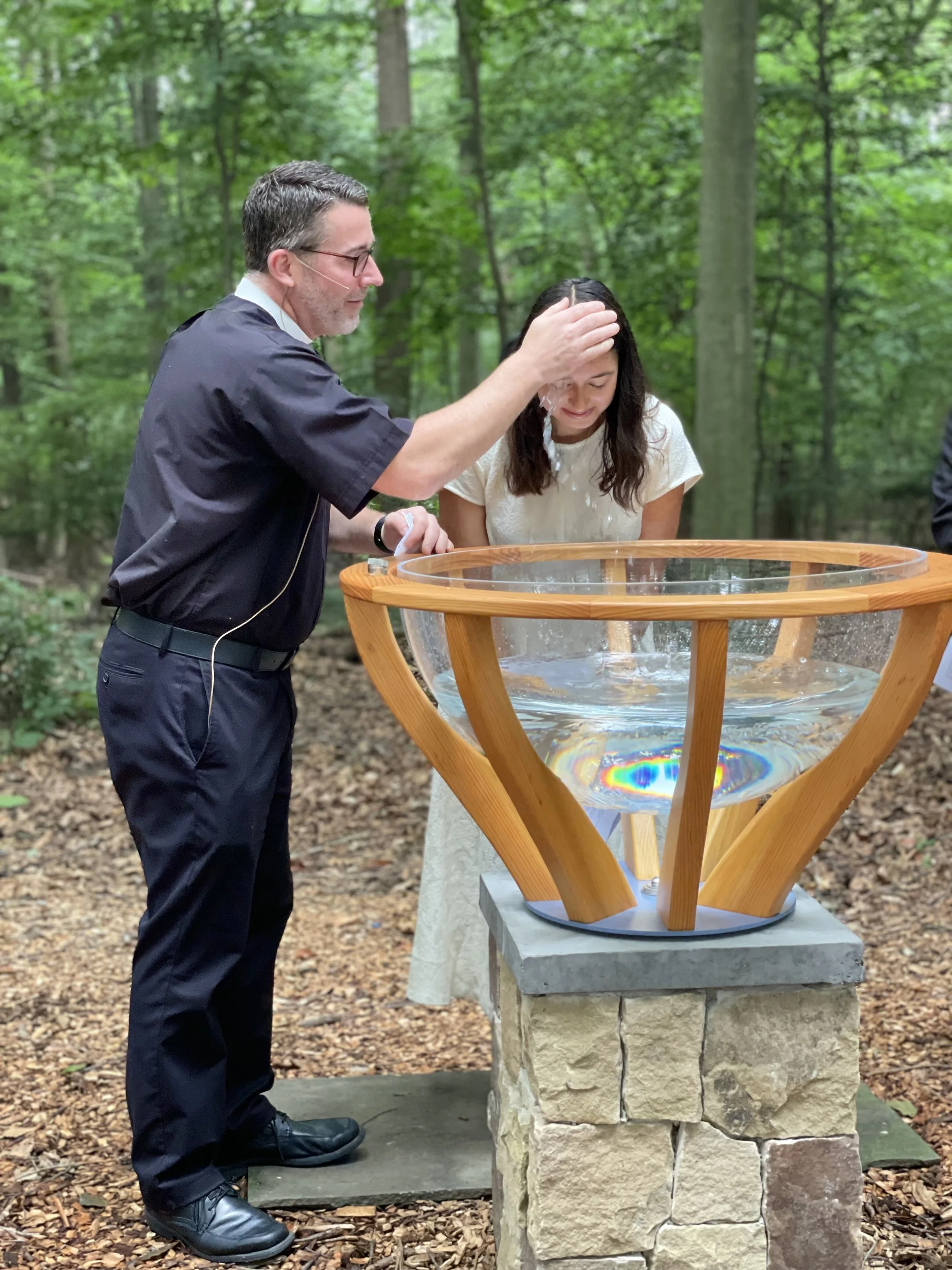 A man in black clothes baptizing a woman in a white dress outdoors in a wooded area. The man is pouring water over the woman's head as she bows her head with her eyes closed.