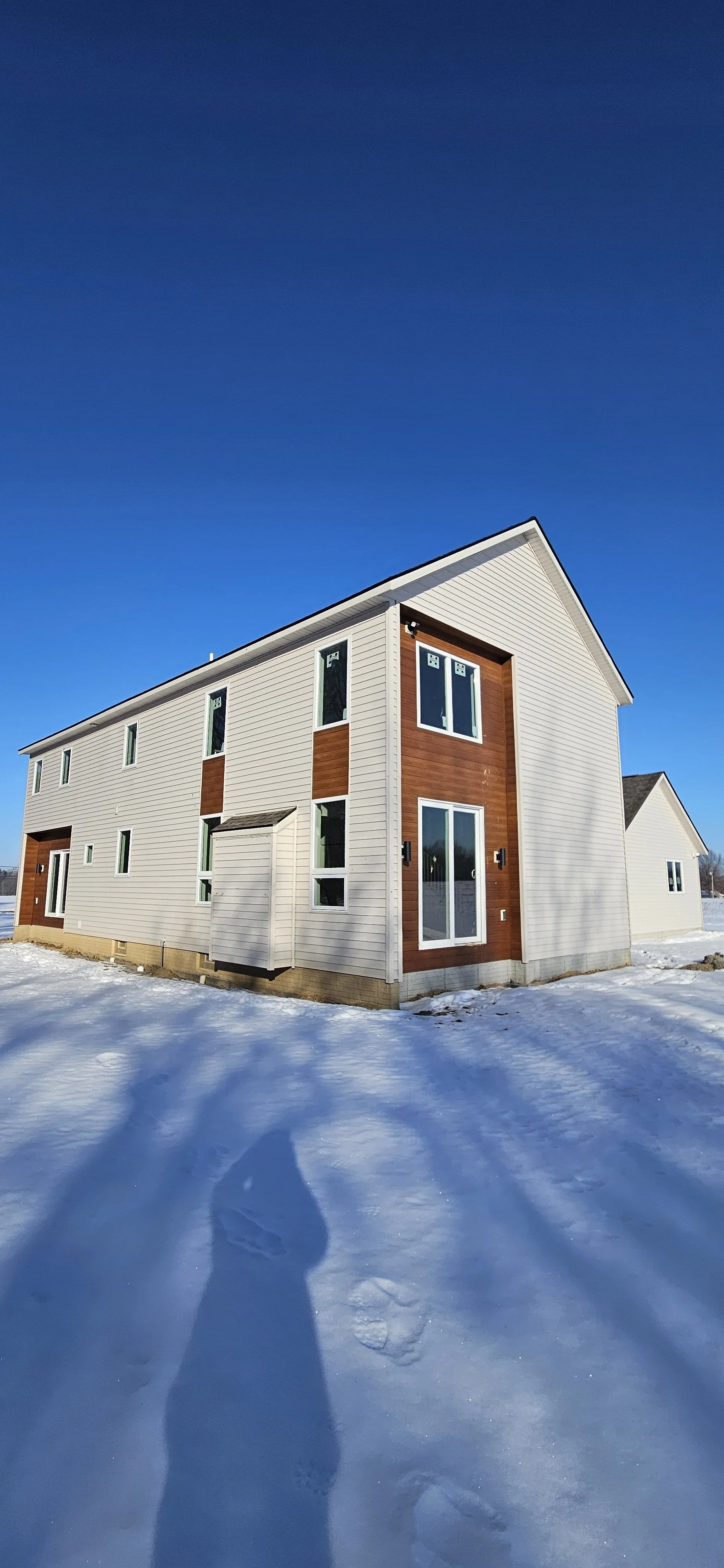 Newly constructed multi-story house with white siding and brown accents, situated in a snowy landscape under a clear blue sky.