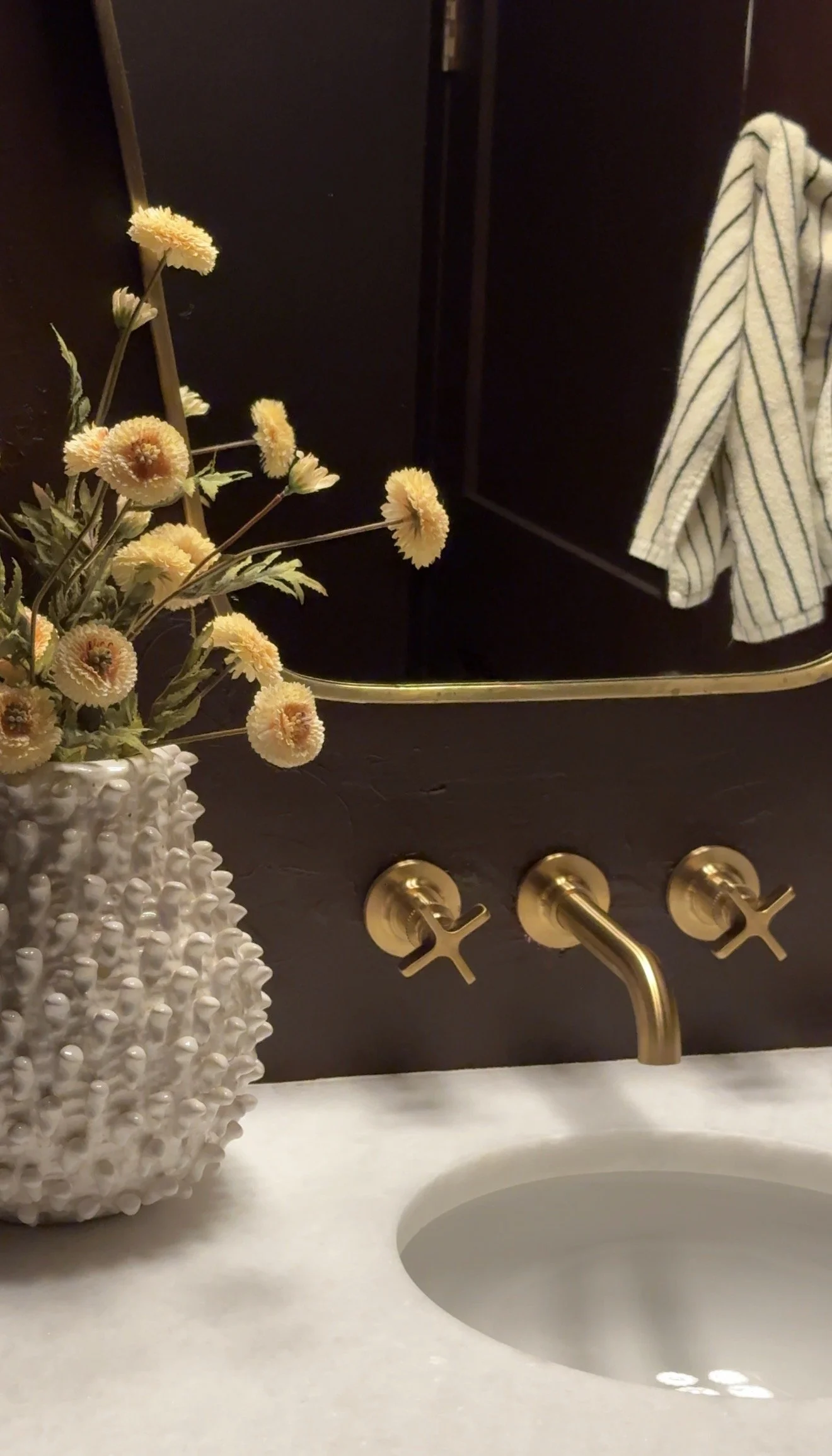 A bathroom sink with a white countertop, a floral vase with white textured surface holding yellow flowers, and a dark wall with a gold-colored faucet and hooks. A striped towel is hanging on the right side of the image.