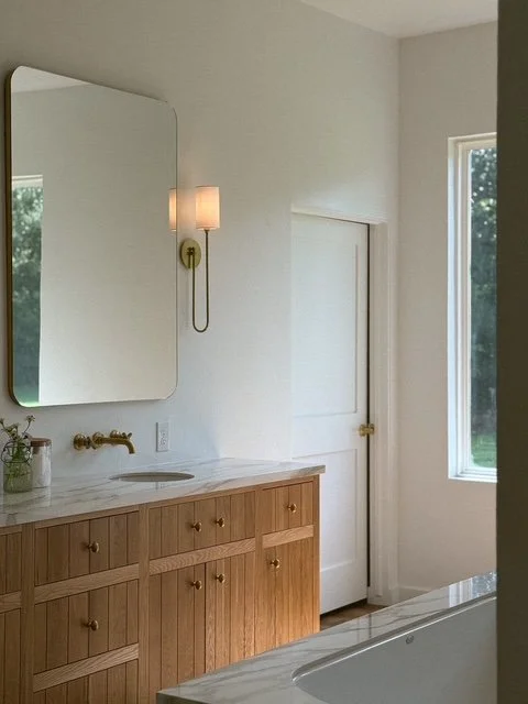 A bathroom with a wooden vanity, a wall-mounted mirror, a wall lamp, a door, and a window with greenery outside.