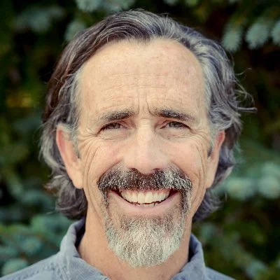 A smiling middle-aged man with gray hair, beard, and mustache, standing outdoors with blurred greenery in the background.