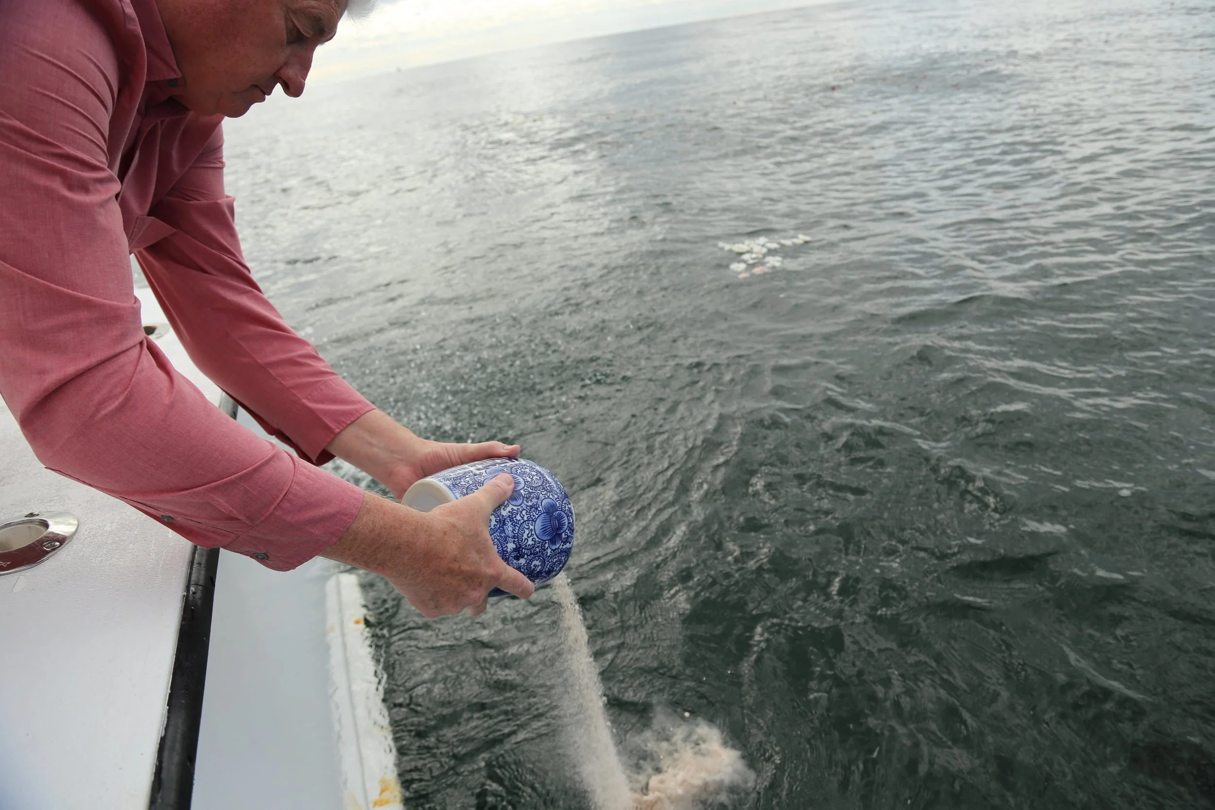 A person in a pink shirt is holding a blue and white patterned cup over the edge of a boat, pouring liquid into the ocean. The ocean water is visible with gentle waves, and the sky is cloudy.