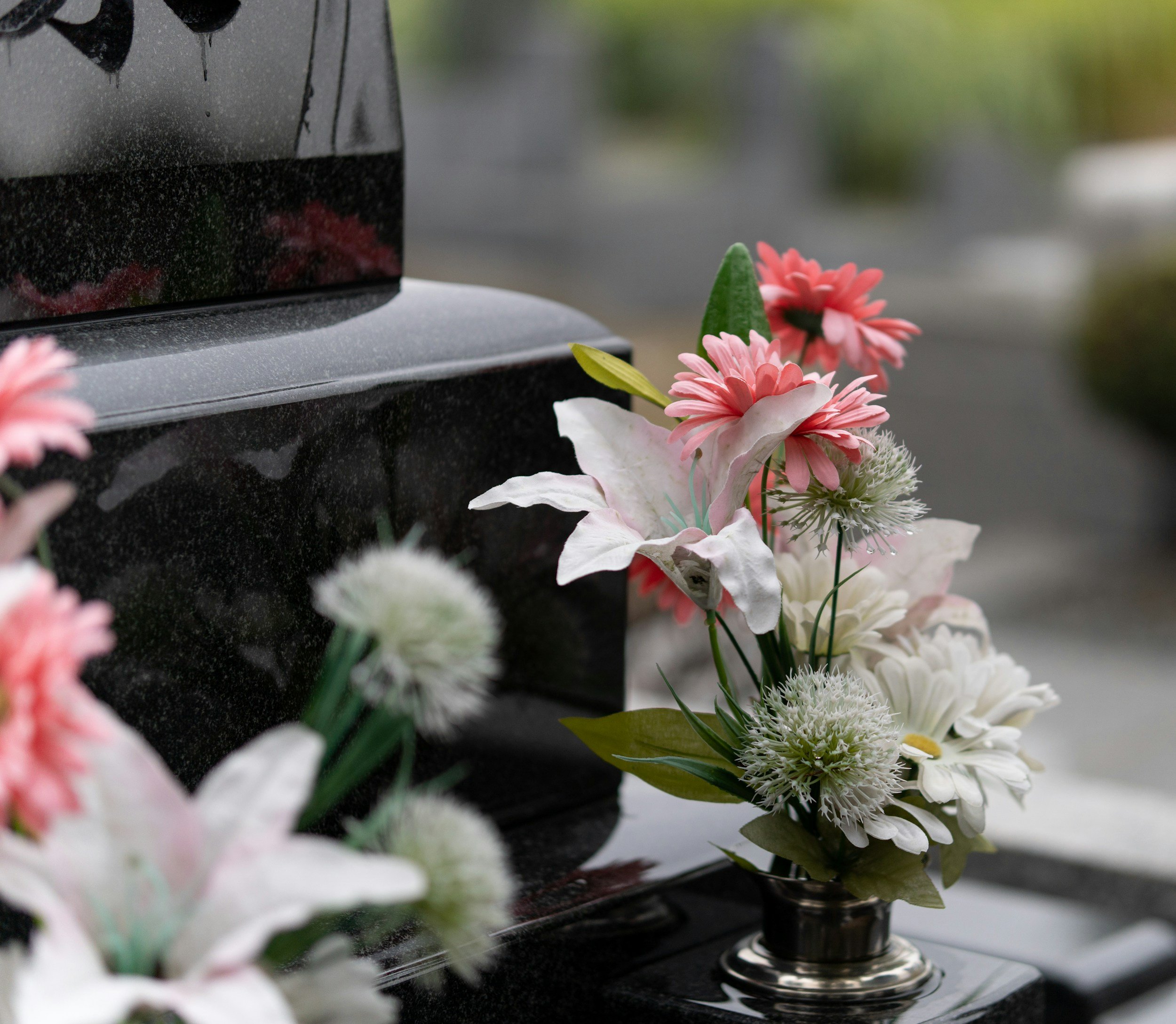 A funeral urn with pink, white, and green floral arrangements around it, placed on a black surface with a blurred outdoor background.