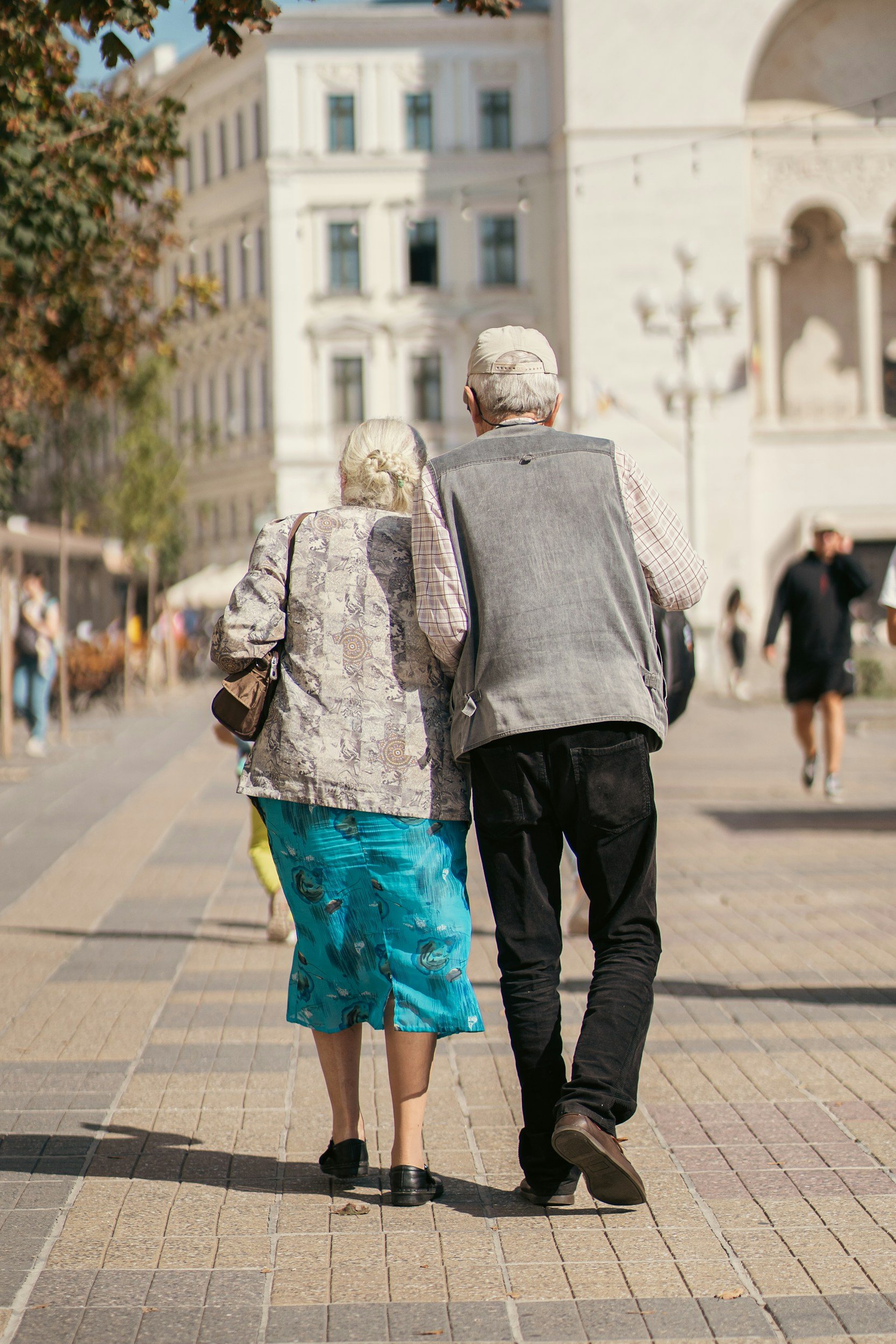 An elderly man and woman walking together down a city street, seen from behind, with large white buildings and other pedestrians in the background.