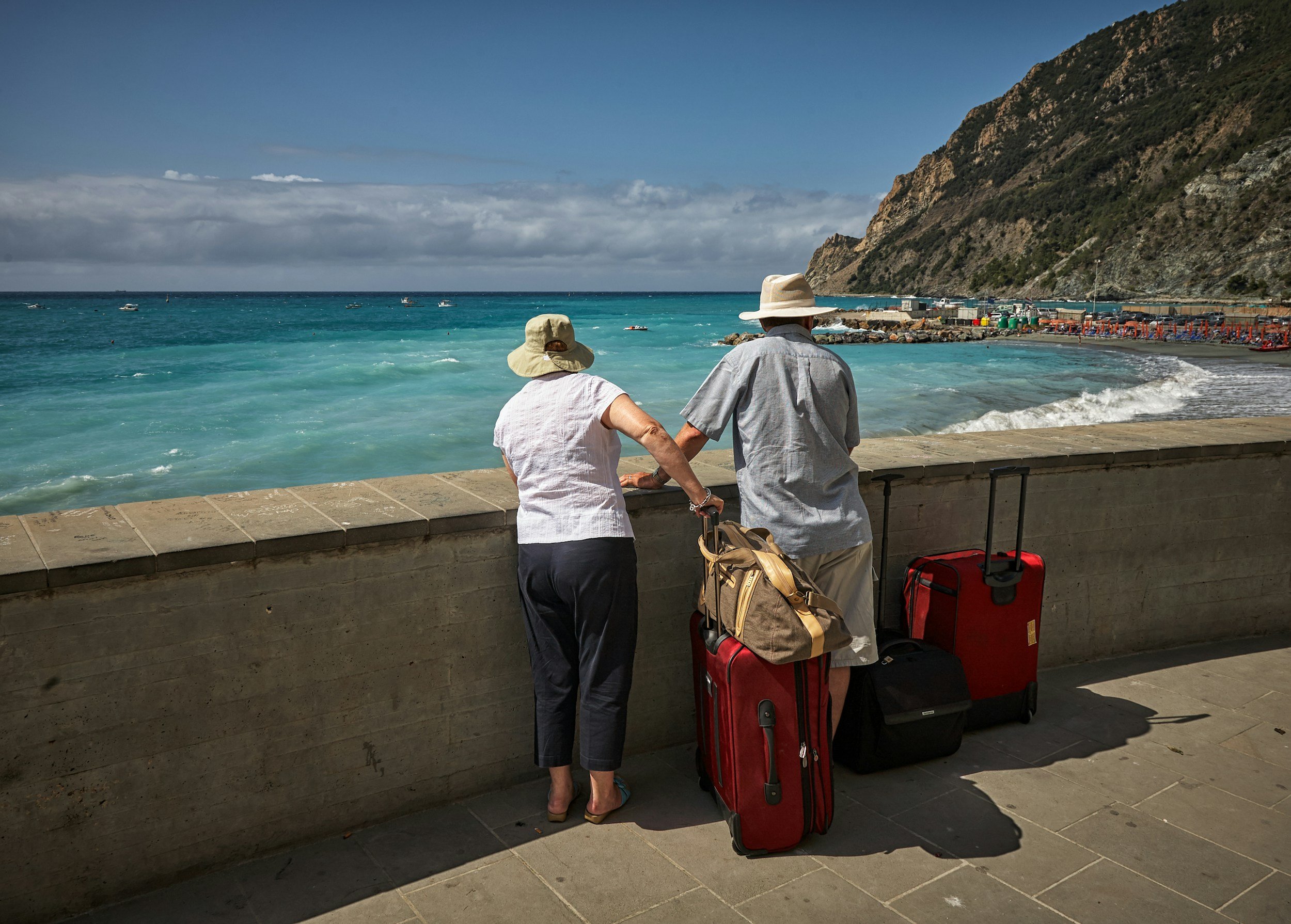 An elderly couple stands by a stone barrier looking out at the ocean, with luggage beside them, in a coastal area with a mountain on the right and several small boats in the water.