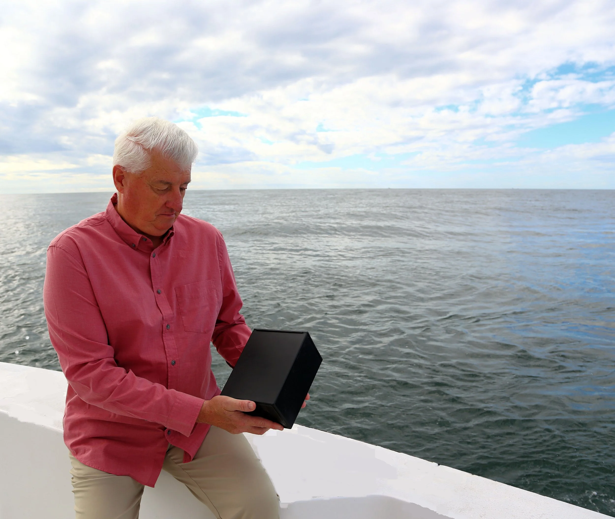 An older man with white hair wearing a pink shirt and beige pants holding a laptop on the deck of a boat, with open water and a cloudy sky in the background.