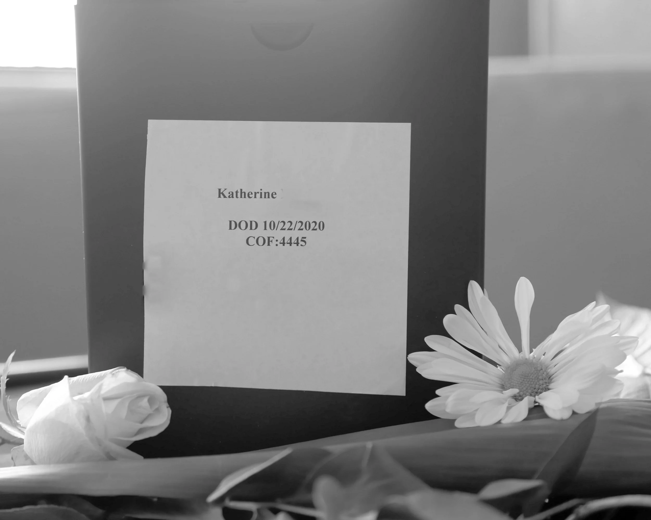 A black-and-white photo of a cardboard box with a label that reads 'Katherine DOD 10/22/2020 COF:4445,' surrounded by flowers.