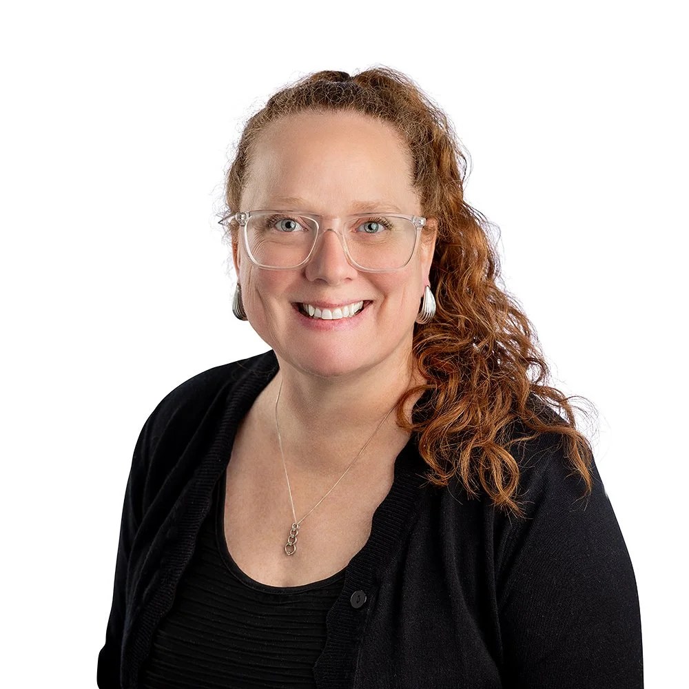 Portrait of a smiling woman with red hair in a ponytail. She's wearing glasses and a black top.