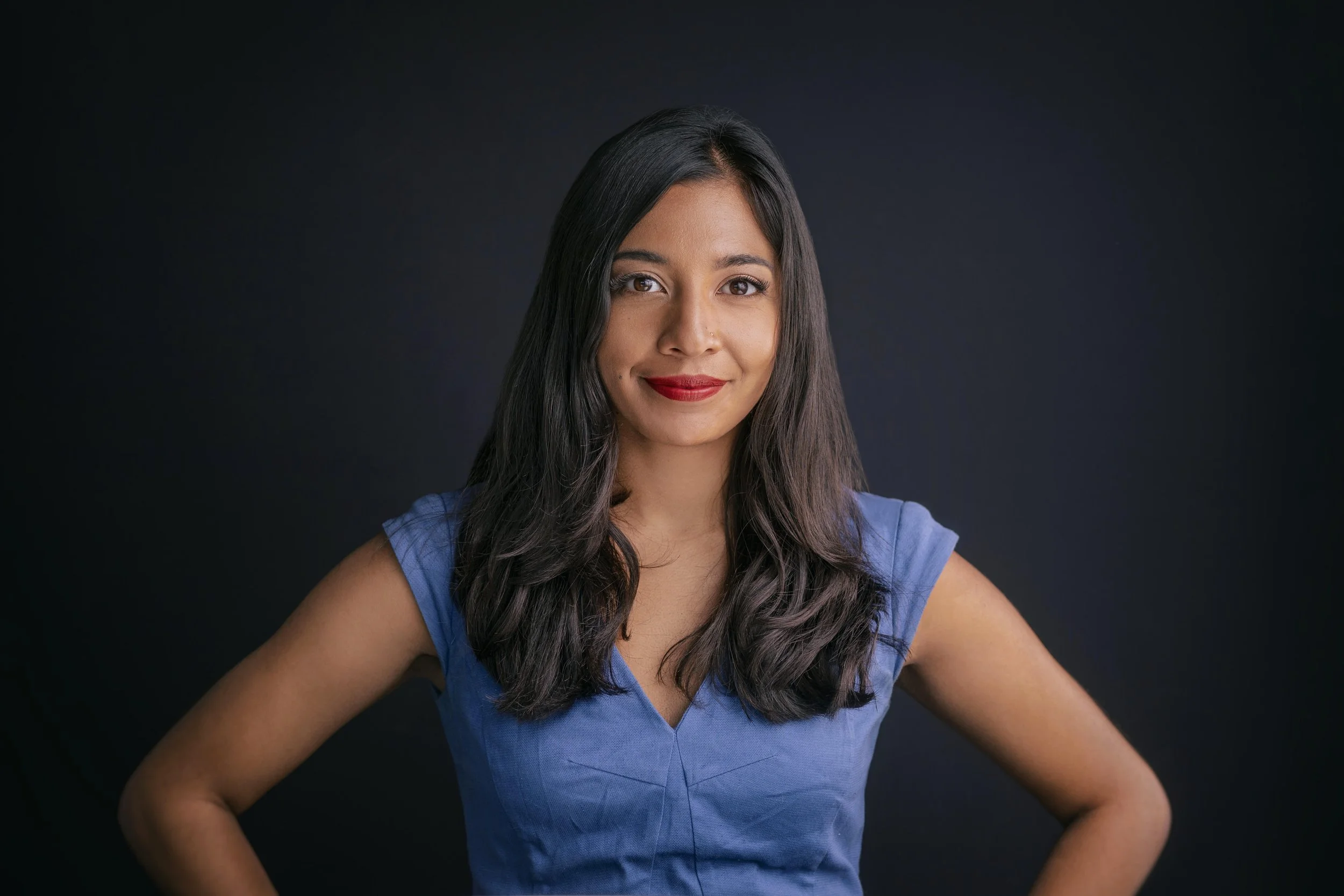 A woman with long dark hair, red lipstick, and wearing a blue sleeveless top, smiling slightly and posing against a dark background.