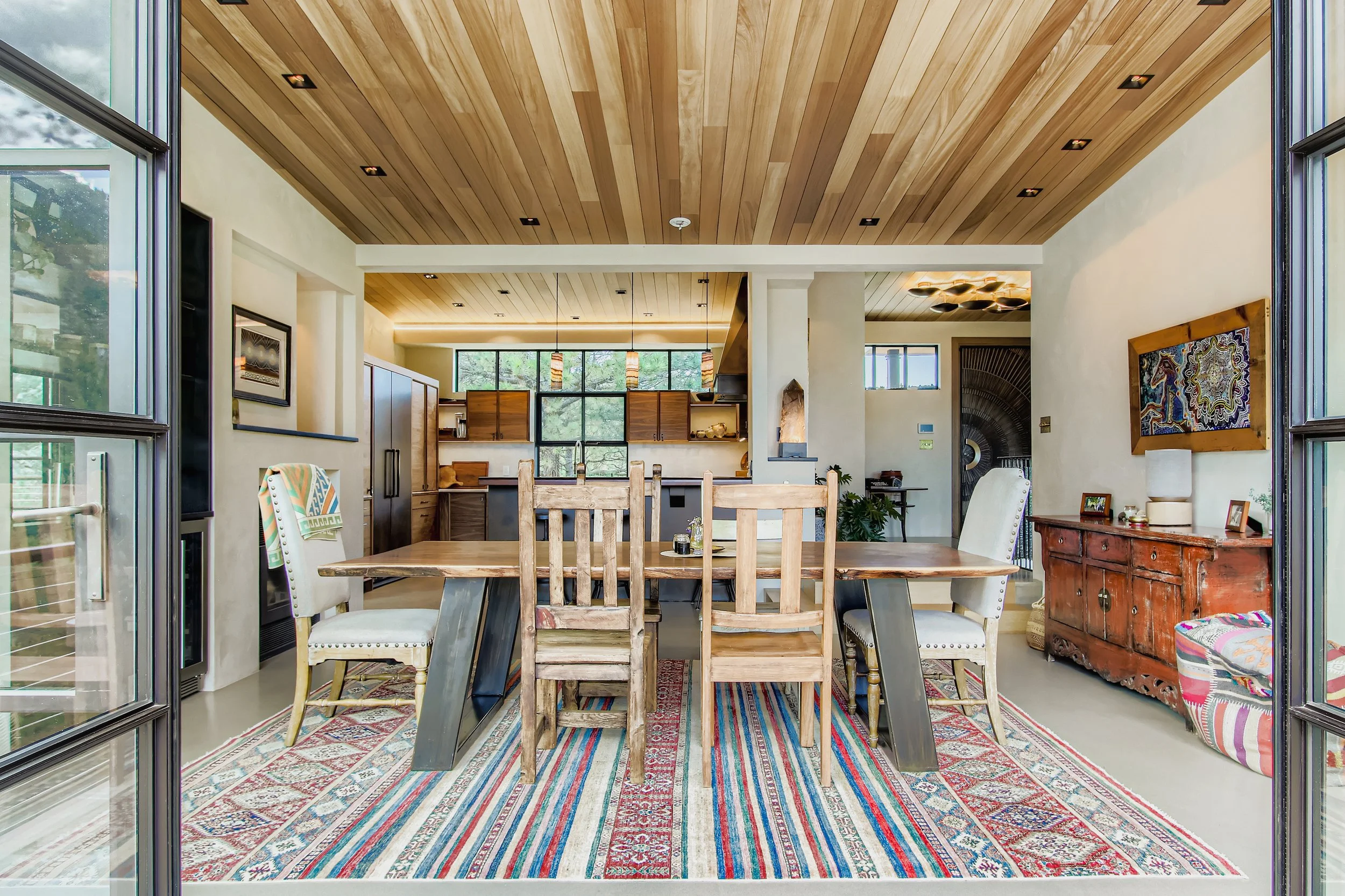 Dining room at Onyx Ridge, Boulder County, CO, with wooden ceiling, colorful striped rug, wooden and upholstered chairs around a wood table, and open kitchen with large windows and pendant lighting.