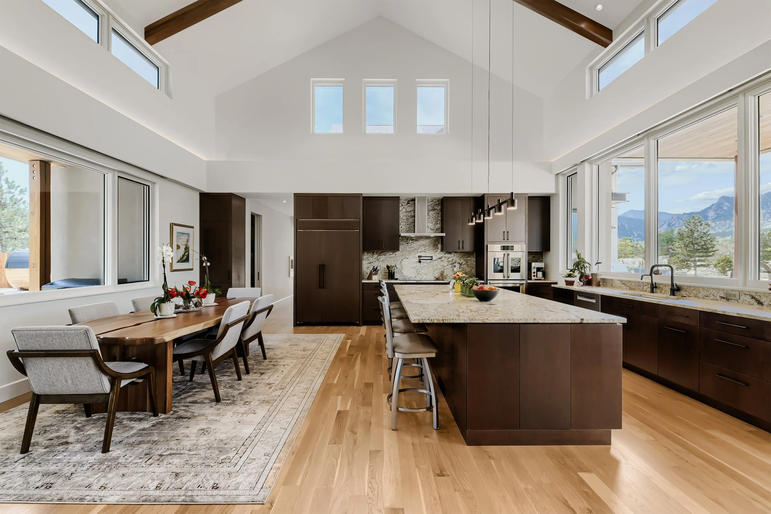 Open-concept kitchen and dining area with vaulted ceilings, exposed wood beams, large island, and expansive windows overlooking the Boulder landscape.