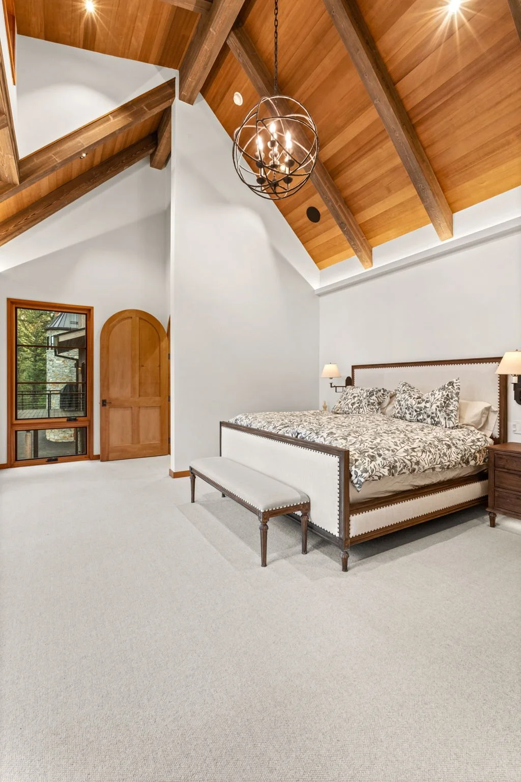 Bedroom with vaulted wood ceiling, chandelier, large bed with floral bedding, and arched doorway with window.