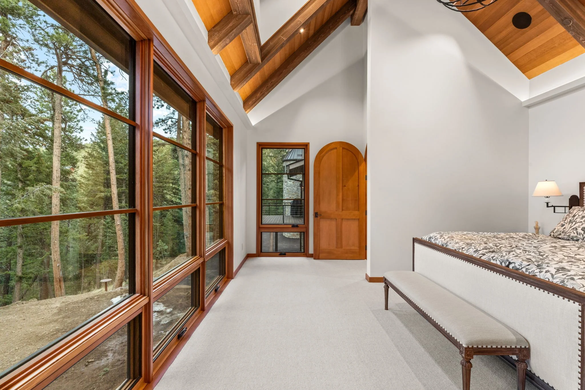 Bedroom with large window overlooking trees, arched wooden door, and bed with floral bedding.