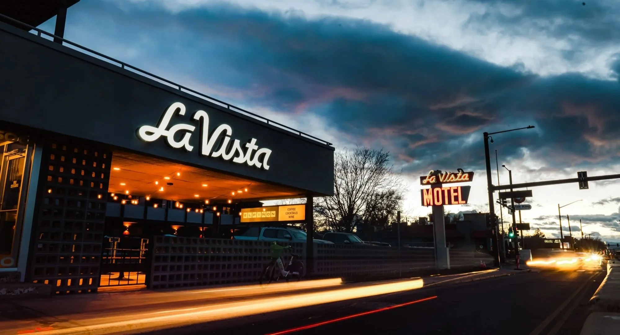La Vista Motel in Denver, CO at dusk, showing mid-century modern exterior with illuminated signage, outdoor seating under string lights, and light trails from passing vehicles.
-Designed by Sopher Sparn Architects