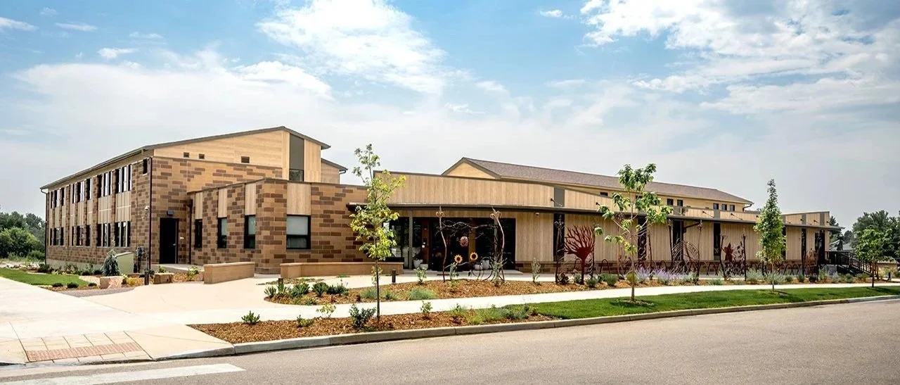 Exterior view of Shining Mountain Waldorf School High School in Boulder, CO, showing a modern multi-story building with tan brick and wood facade, large windows, landscaped front yard, and a clear sky.
-Designed by Sopher Sparn Architects