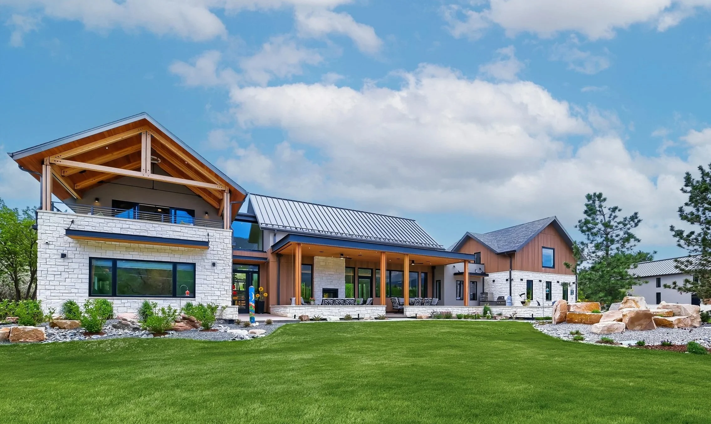 Modern mountain residence with stone and wood exterior, covered outdoor living space, metal roof accents, and expansive lawn set against a bright blue sky. Designed by Sopher Sparn Architects