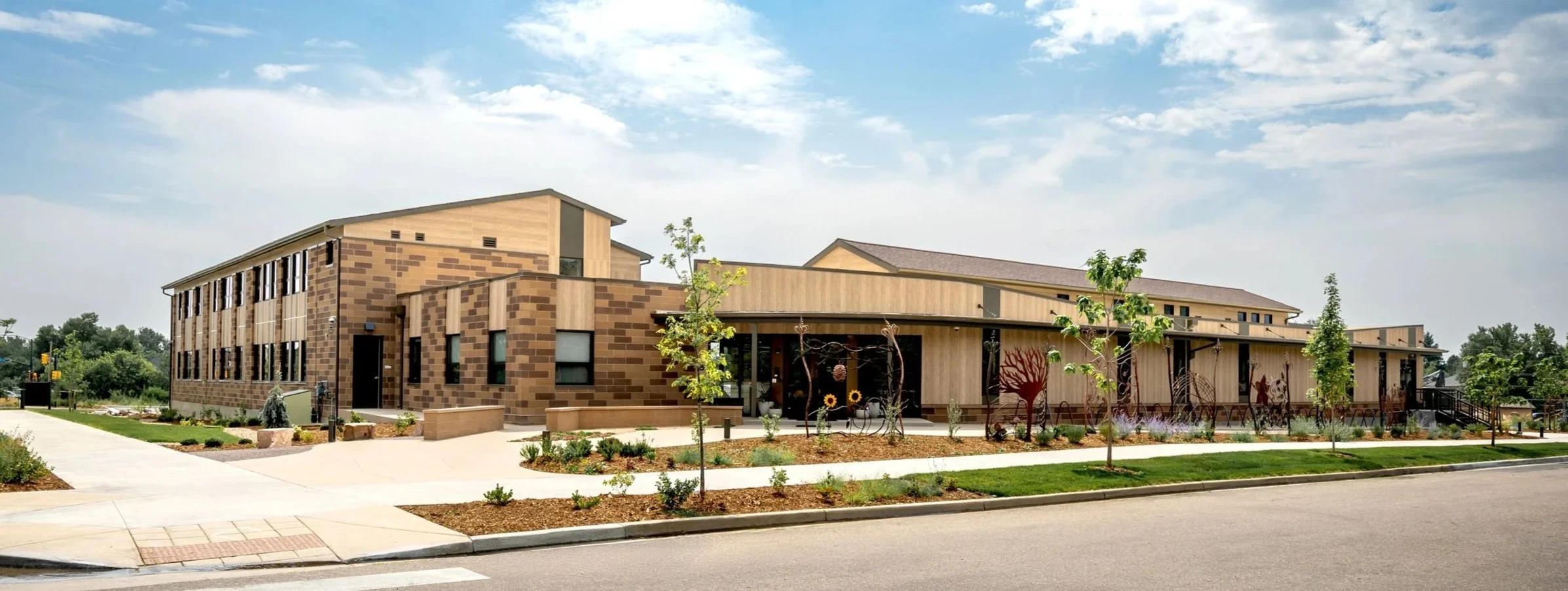Exterior view of Shining Mountain Waldorf School High School in Boulder, CO, showing a modern multi-story building with tan brick and wood facade, large windows, landscaped front yard, and a clear sky.
-Designed by Sopher Sparn Architects