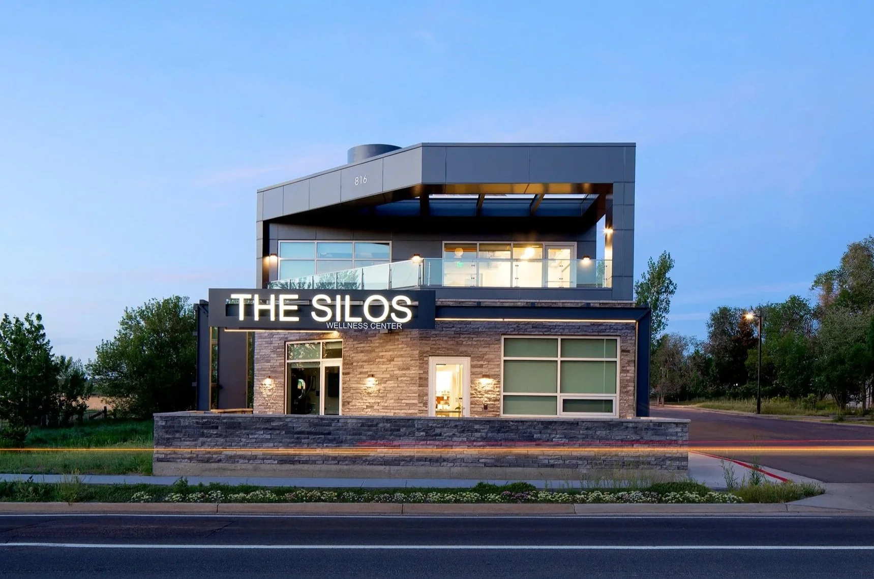 The Silos Wellness Center in Lafayette, CO, modern two-story building with metal, stone, and glass exterior, outdoor yoga patio, and illuminated signage along the street.
-Designed by Sopher Sparn Architects