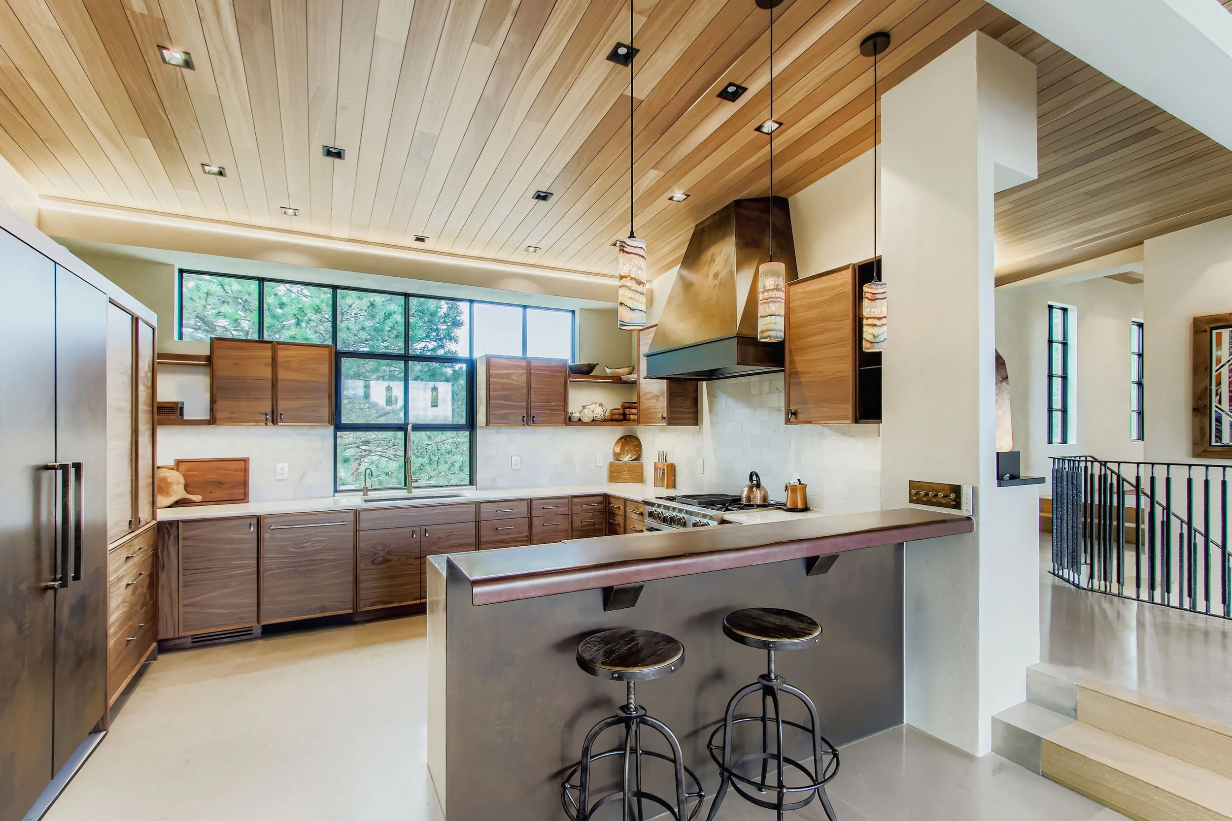 Kitchen in Onyx Ridge, Boulder County, CO, with wooden cabinets, a large window, stainless steel refrigerator, range with hood, hanging pendant lights, and a breakfast bar with two wooden stools.