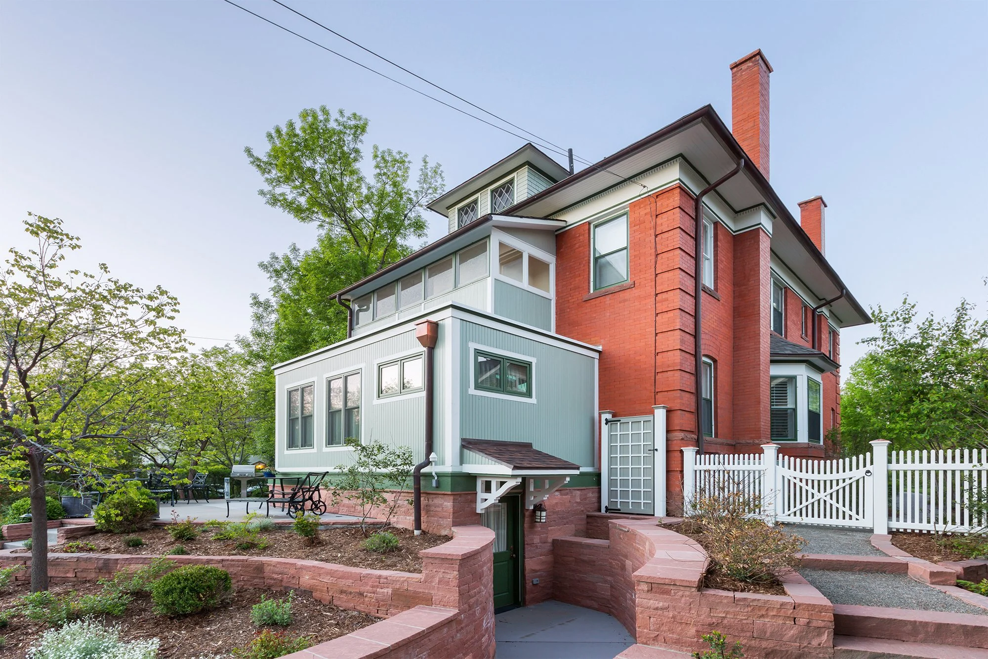 Backyard patio with a round table and six chairs, surrounded by shrubs and trees with string lights, adjacent to a multi-story house with green and red brick exterior walls, windows, and a barbecue grill on the patio.