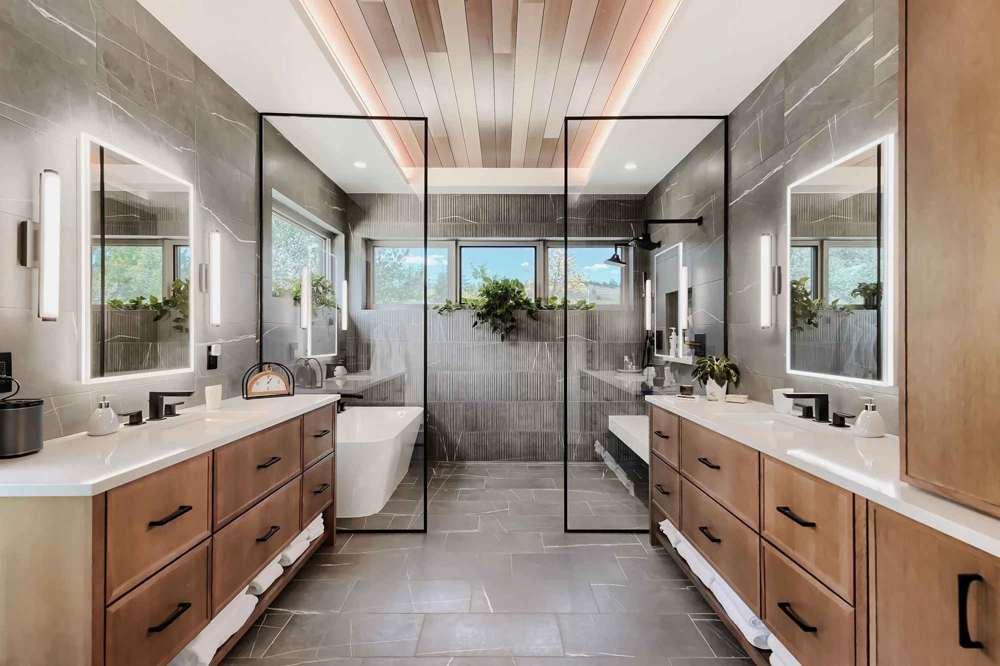 Bright modern bathroom with dual wood vanities, large mirrors, skylight, and walk-in shower with natural light.