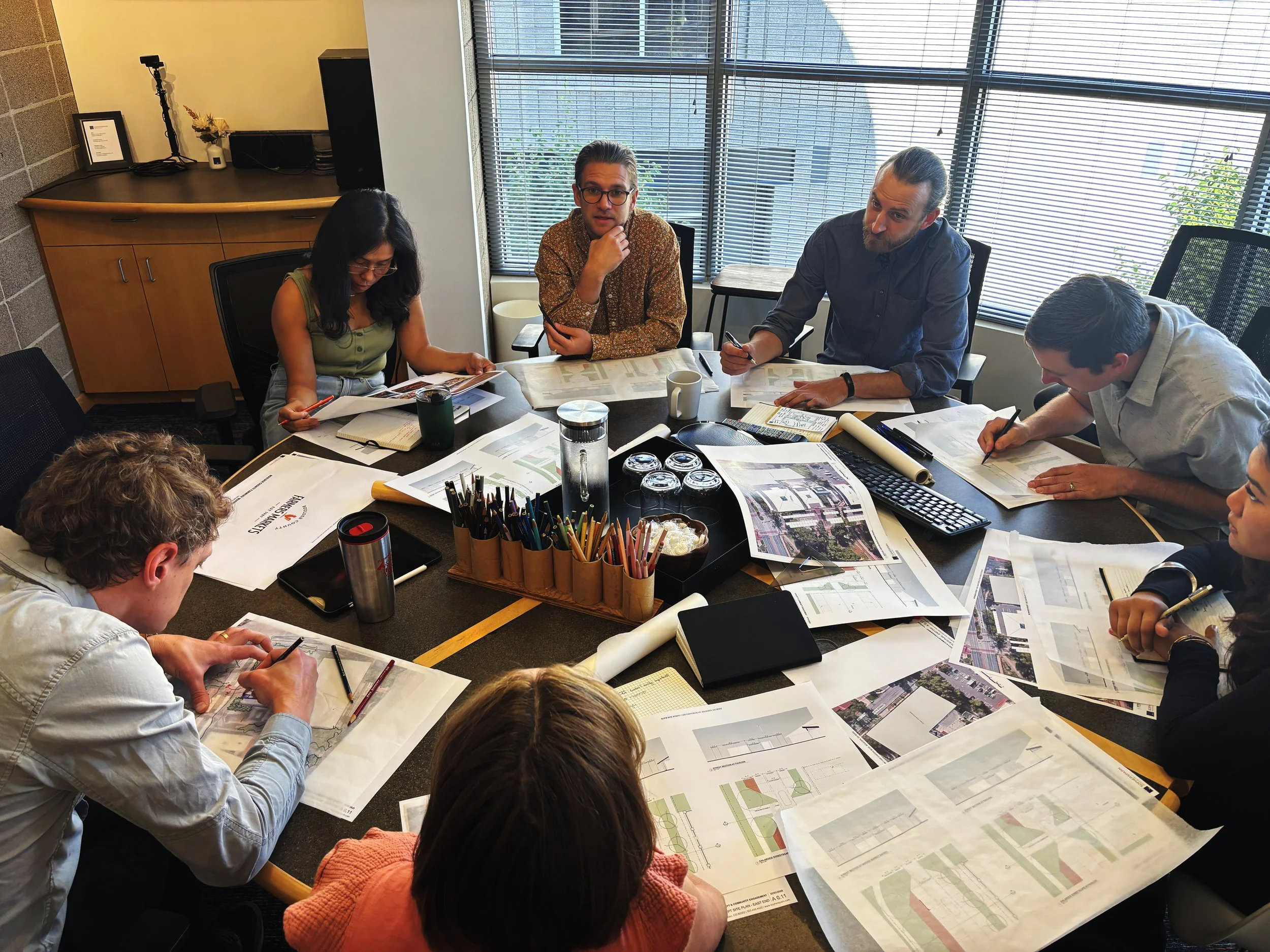 A group of six people sitting around a conference table covered with architectural plans and documents, engaged in a discussion in a modern office with large windows and blinds.