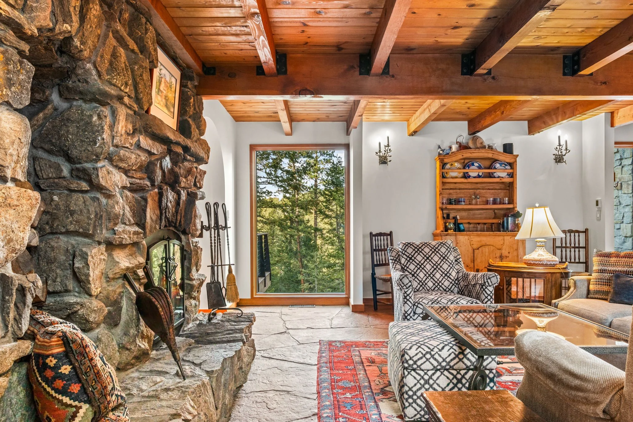 Living room at the Cedar & Stone residence in Jefferson County, Colorado, with stone fireplace, exposed wood ceiling beams, and large windows overlooking the forest.