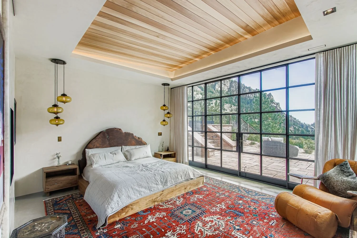 Bedroom at Onyx Ridge, Boulder County, CO, with large bed and wooden headboard, nightstands with pendant lights, and floor-to-ceiling window with mountain and patio views.