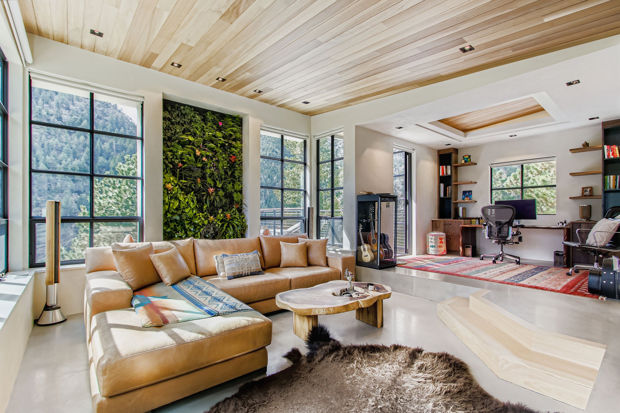 Open-concept living room in Onyx Ridge, Boulder County, CO, featuring large windows, beige leather sectional sofa, wooden coffee table, green living wall, desk with chair, and staircase with rug.