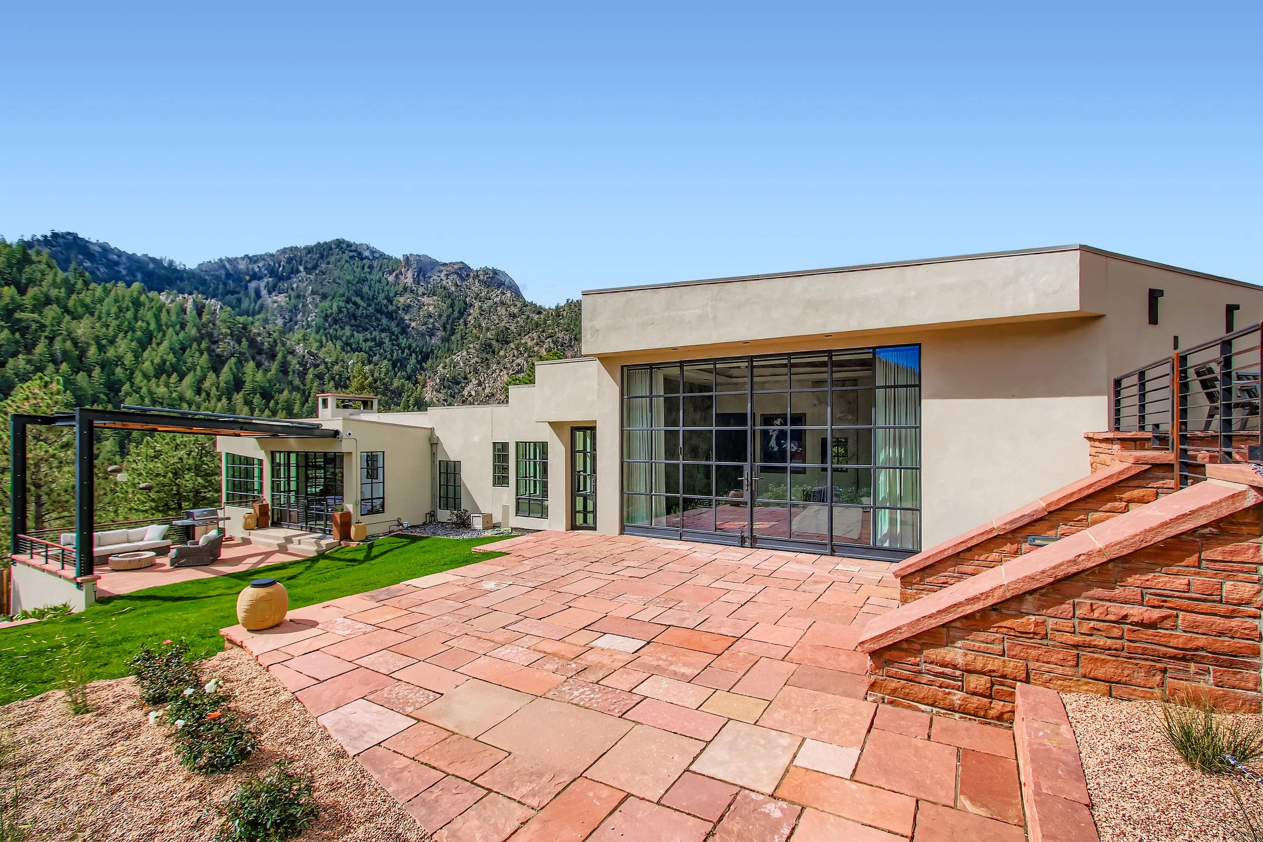 Exterior of Onyx Ridge, Boulder County, CO, with large glass windows, outdoor patio, and hillside landscape with trees and mountains in the background.