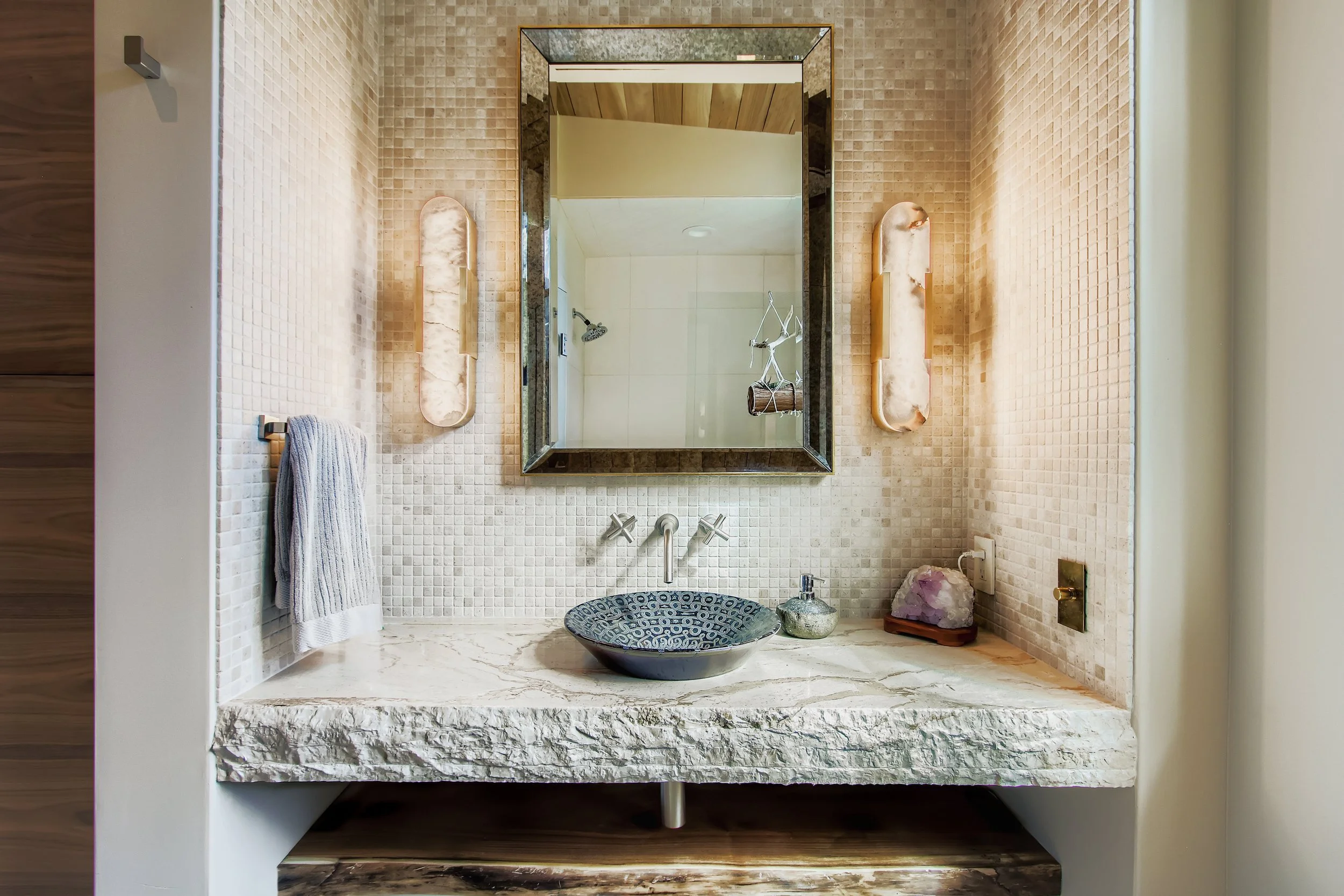 Bathroom at Onyx Ridge, Boulder County, CO, with textured stone countertop, black-and-white vessel sink, wall-mounted faucet, large framed mirror, and decorative accessories on tiled walls.