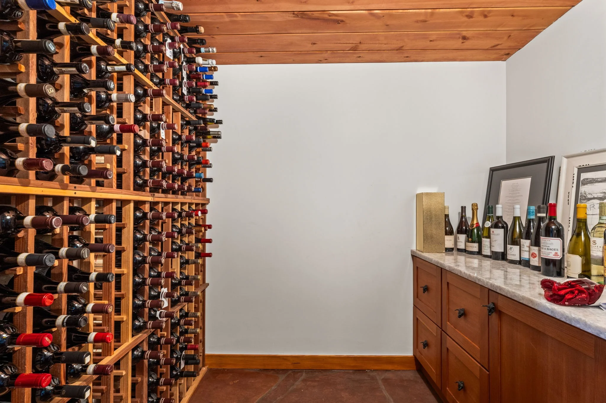 Wine cellar with wooden wine racks filled with bottles and a built-in cabinet displaying additional bottles and framed photos.