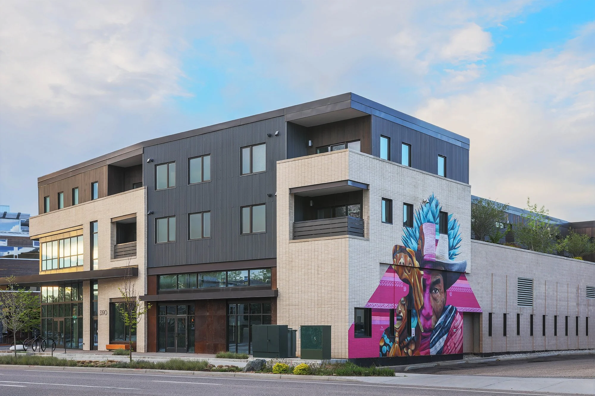 S’PARK Ciclo in Boulder, CO – Colorful mural on the side of a modern multi-story building depicting faces of Indigenous people with traditional accessories, set against a bright pink background with stylized plants and animals.