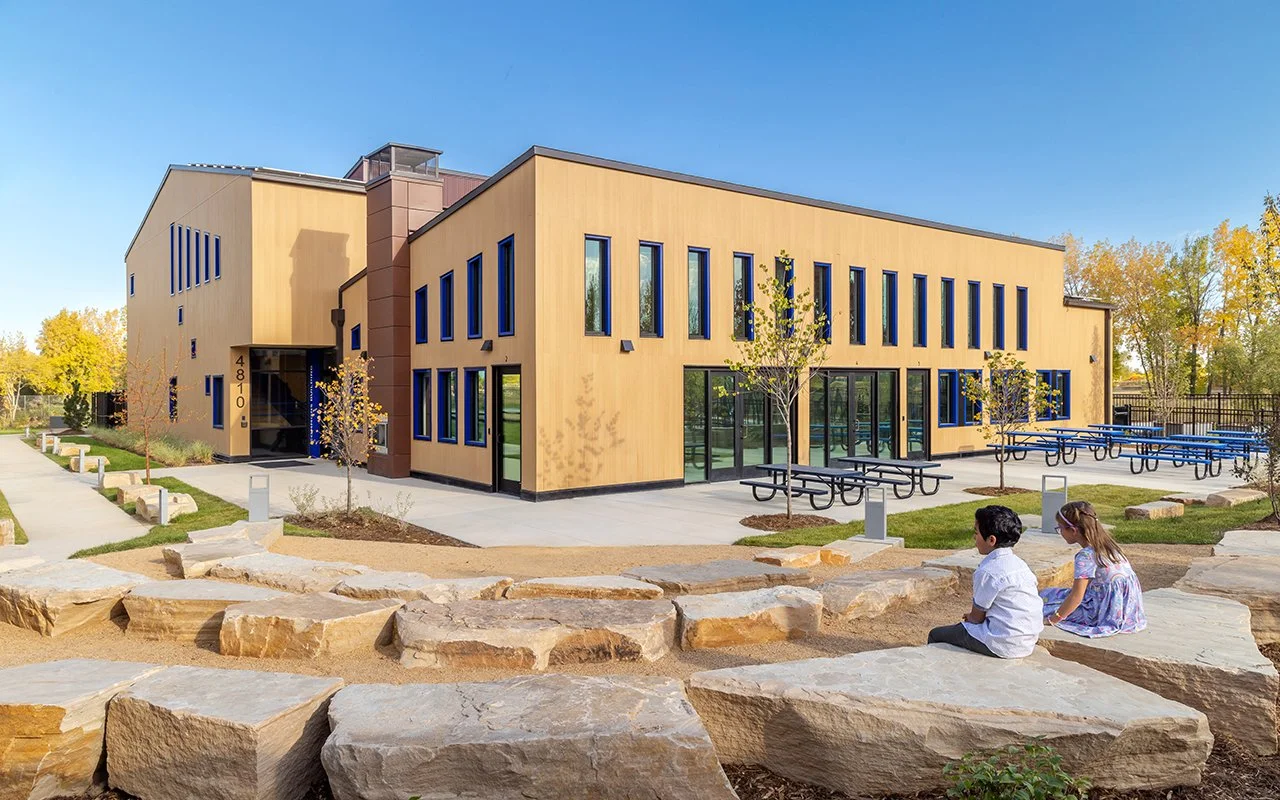 Exterior angle of Boulder Country Day School Commons Building showing tan walls, large windows, and landscaped courtyard with rocks and plants.