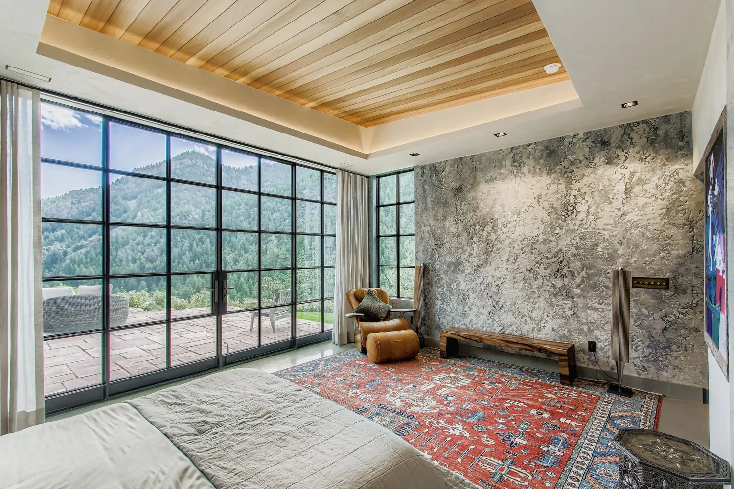 Bedroom at Onyx Ridge, Boulder County, CO, with floor-to-ceiling window showcasing green mountain views, beige bed, orange lounge chair, wooden bench, red patterned rug, textured gray wall, and wooden ceiling.