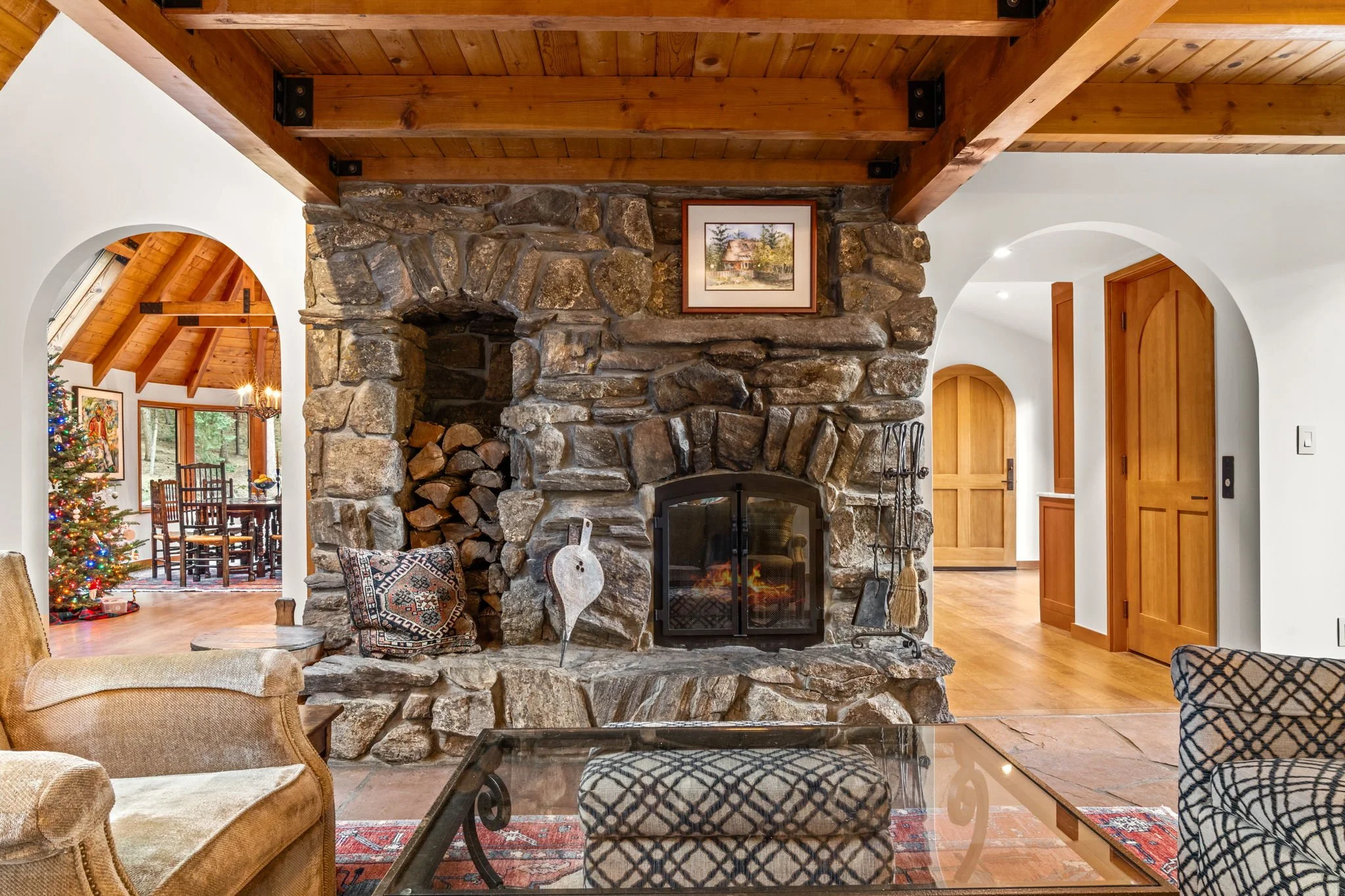 Living room at the Cedar & Stone residence in Jefferson County, Colorado, with stone fireplace, exposed wood ceiling beams, and arched doorway to adjacent room.