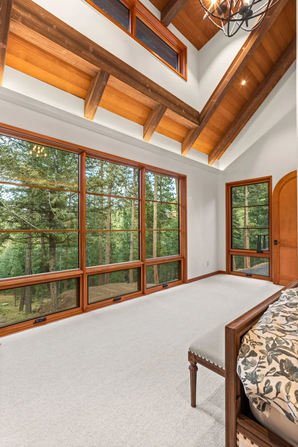 Bedroom at the Cedar & Stone residence in Jefferson County, Colorado, with large windows overlooking the forest, wood ceiling beams, and chandelier.