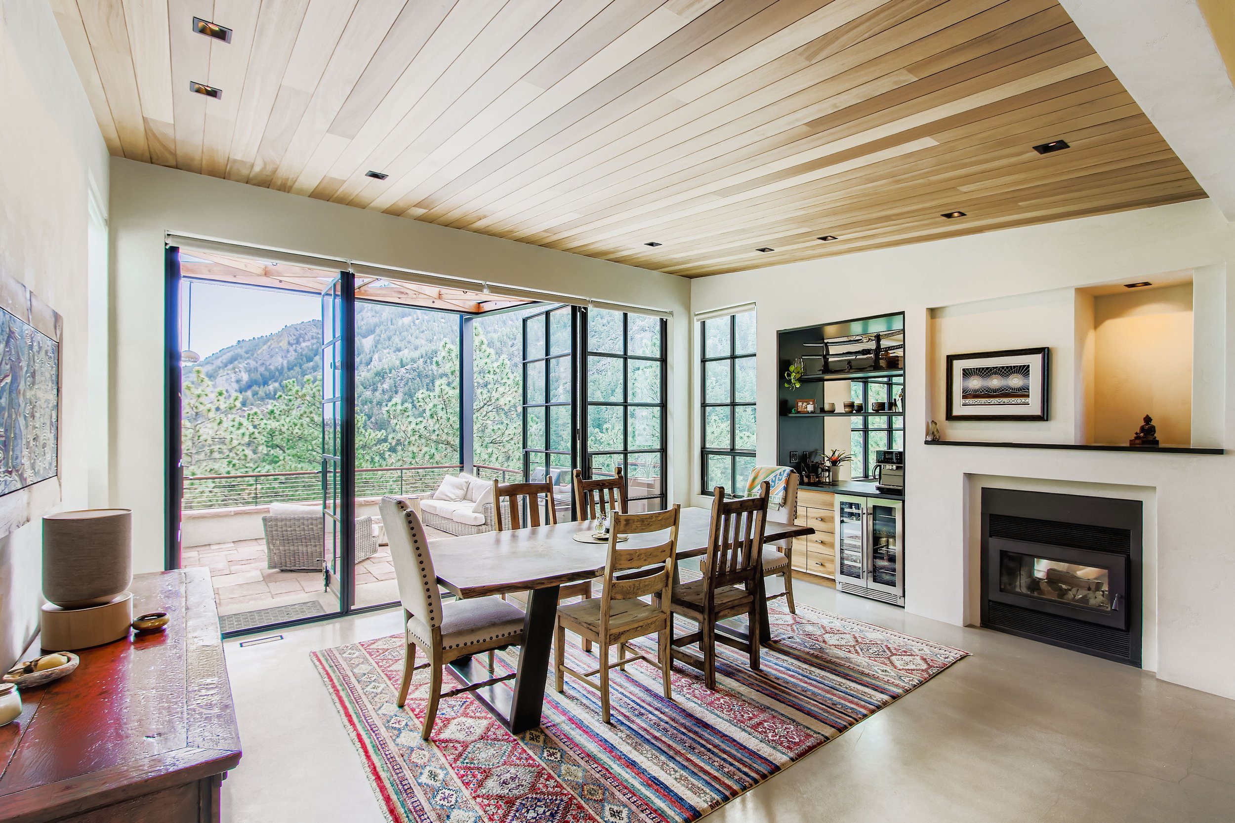 Living and dining area in Onyx Ridge, Boulder County, CO, with large glass doors opening to mountain views, wooden ceiling, fireplace, dining table with chairs, and colorful rug.