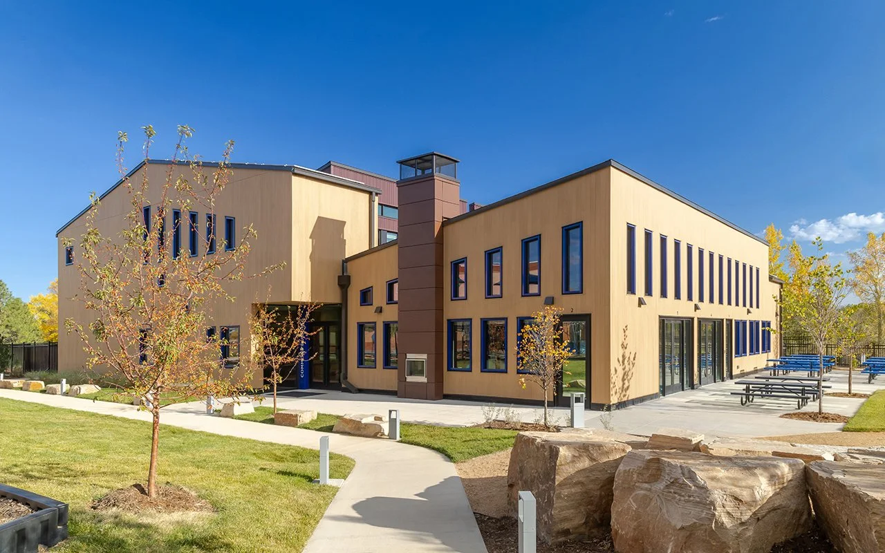 Boulder Country Day School Commons Building in Boulder, CO, showing a modern two-story structure with large windows, tan exterior, outdoor patio with picnic tables, landscaped yard, and trees under a clear blue sky.
-Designed by Sopher Sparn Architec