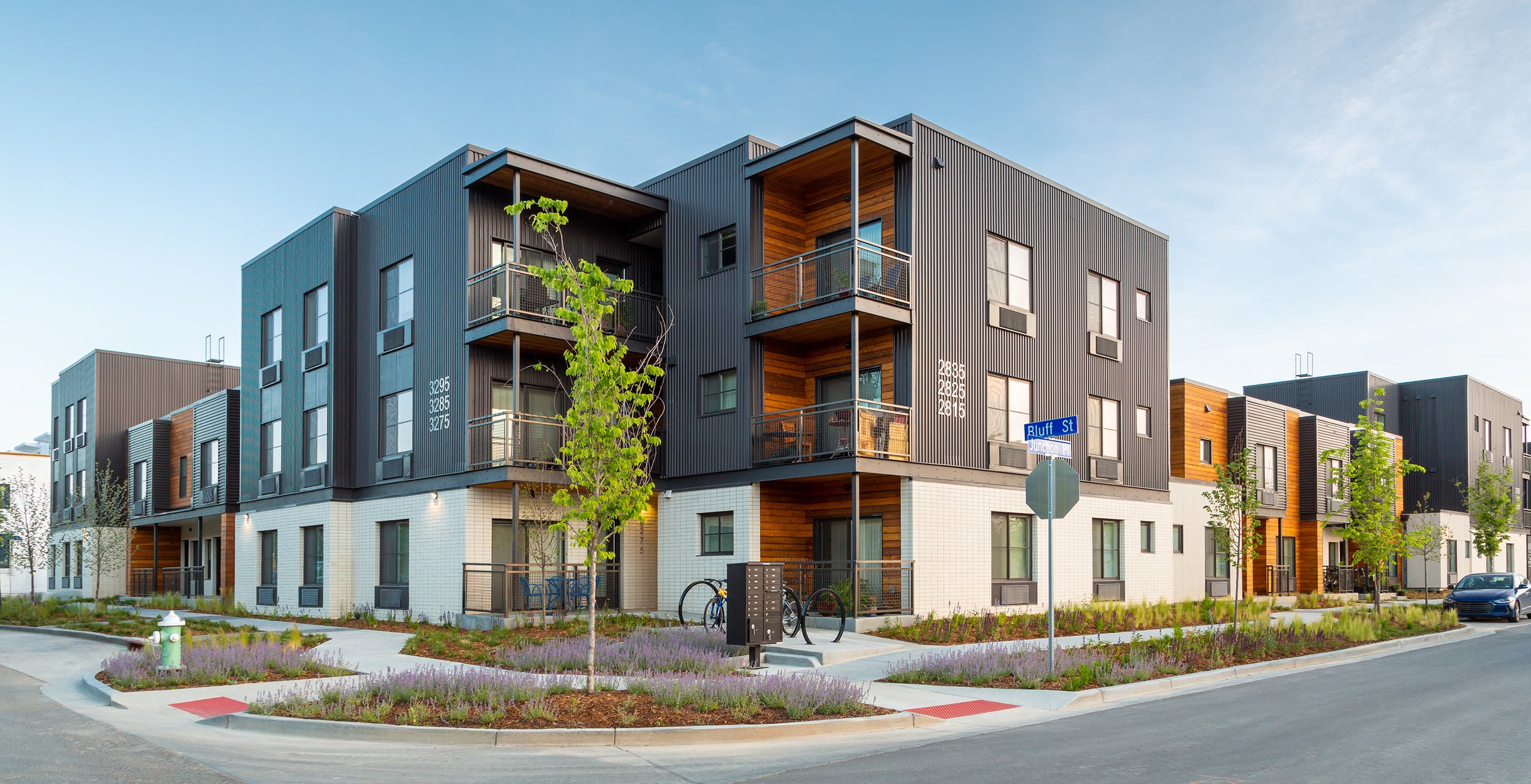 Modern multi-family housing at S’PARK West in Boulder, Colorado, featuring black and wood exterior cladding, private balconies, trees, and bike parking along a residential street. – Designed by Sopher Sparn Architects