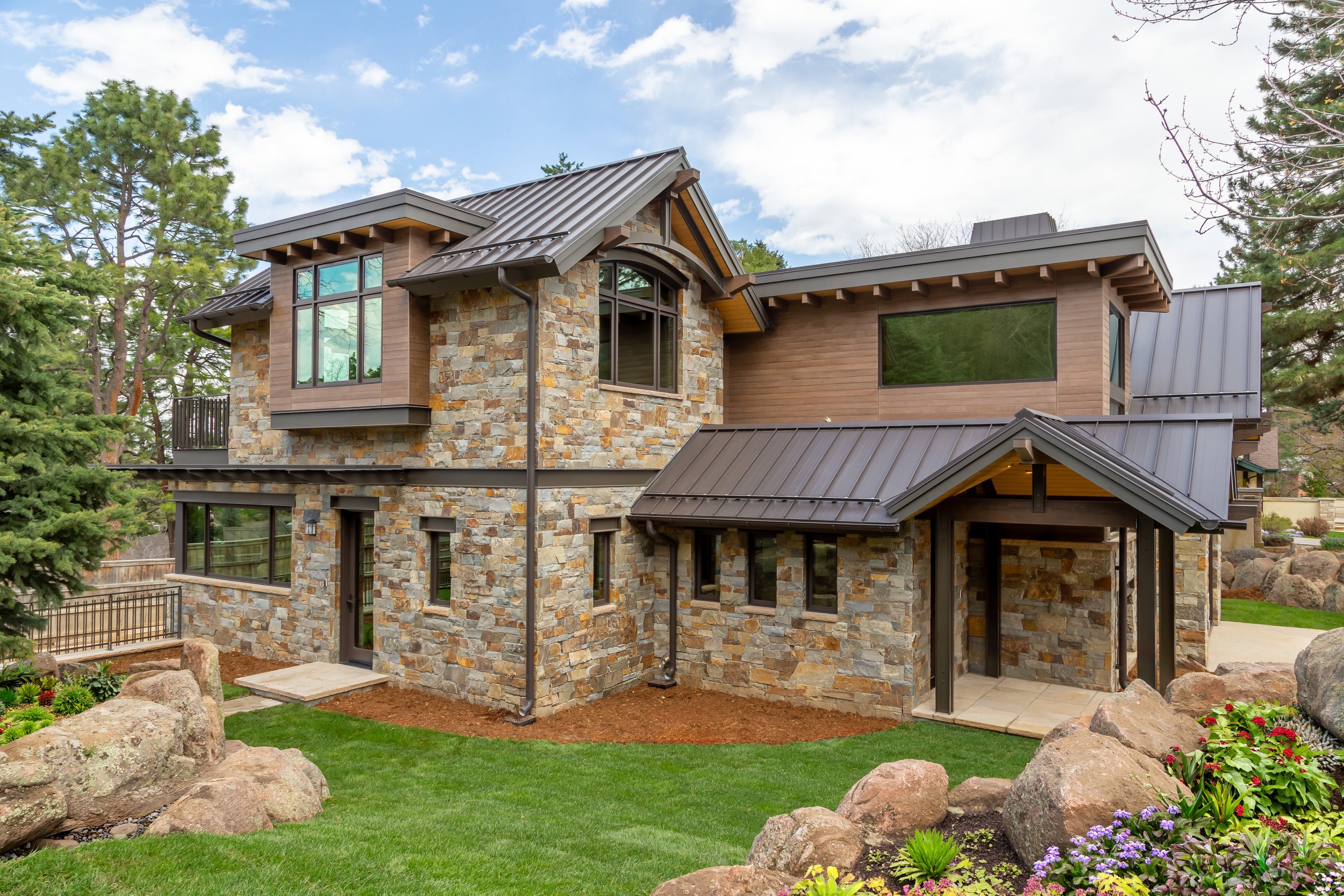 Willowbrook, Boulder, CO – Modern two-story home with stone and wood exterior, metal roof, large windows, and landscaped yard with rocks, flowers, and green grass.