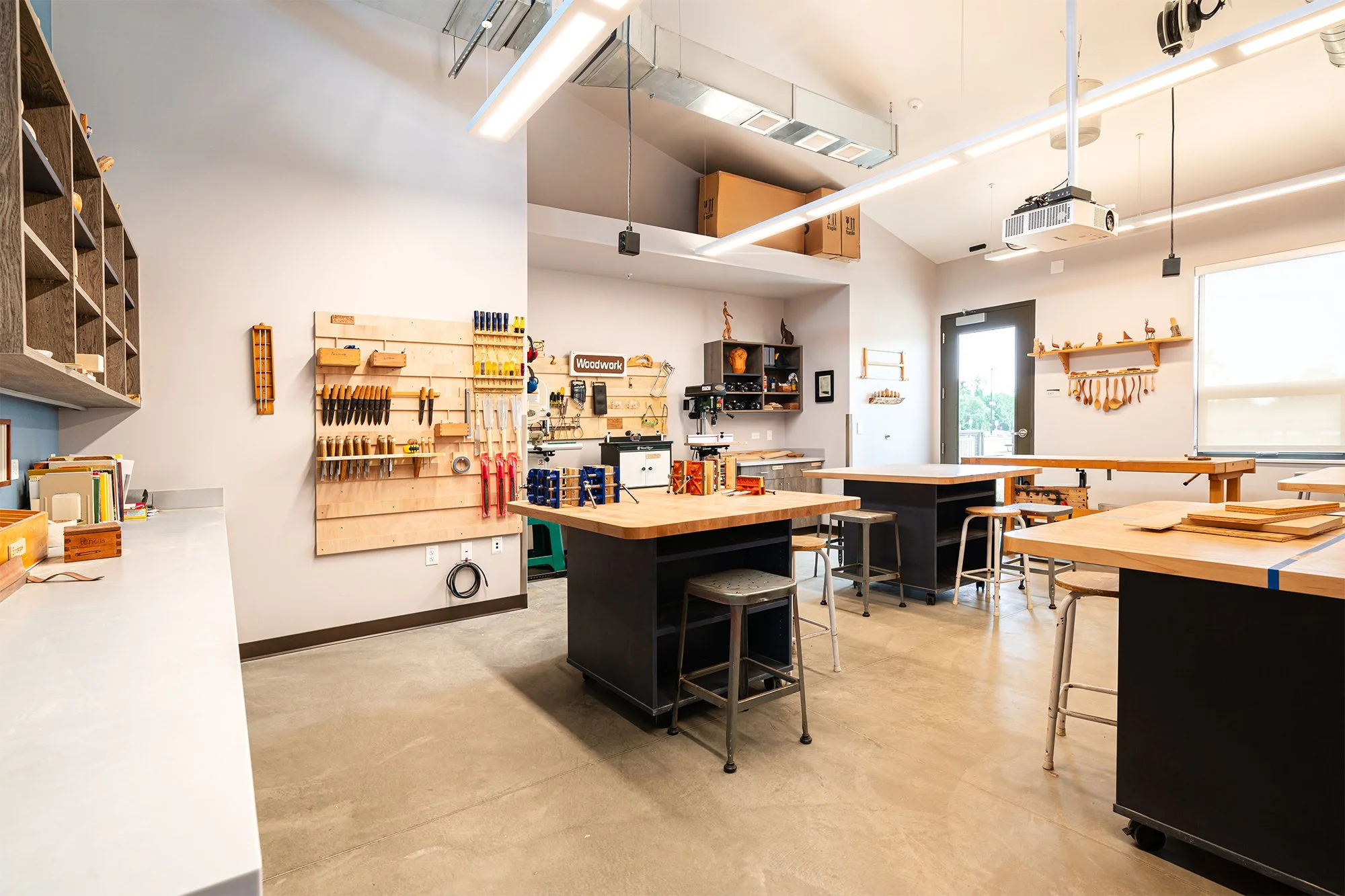 Interior makerspace at Shining Mountain Waldorf School High School in Boulder, CO, featuring worktables, tools, shelves with materials, and high ceilings.