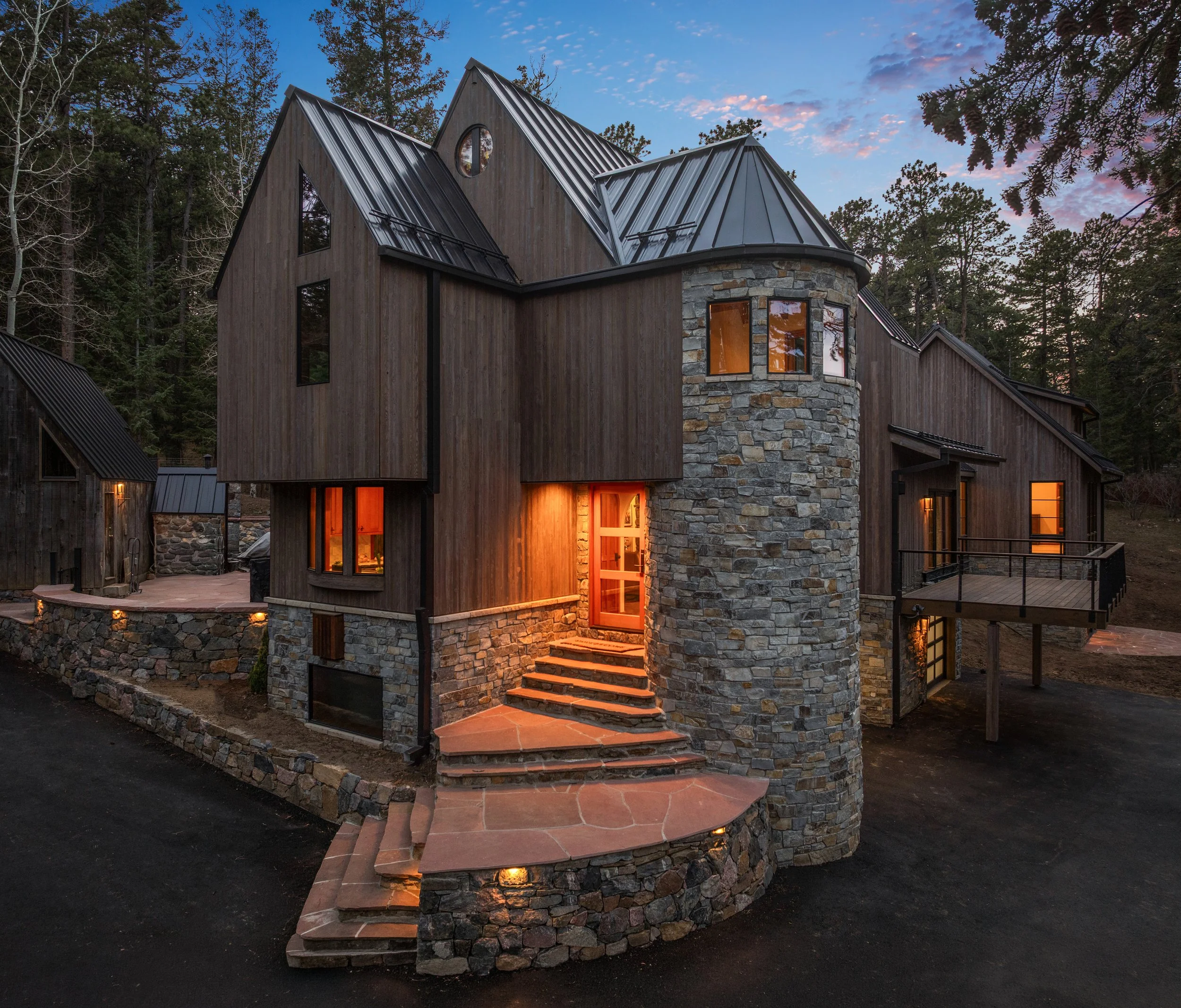Cedar & Stone residence in Jefferson County, Colorado, featuring cedar siding, rounded stone turret, steep metal roofs, and warm interior lighting at dusk.