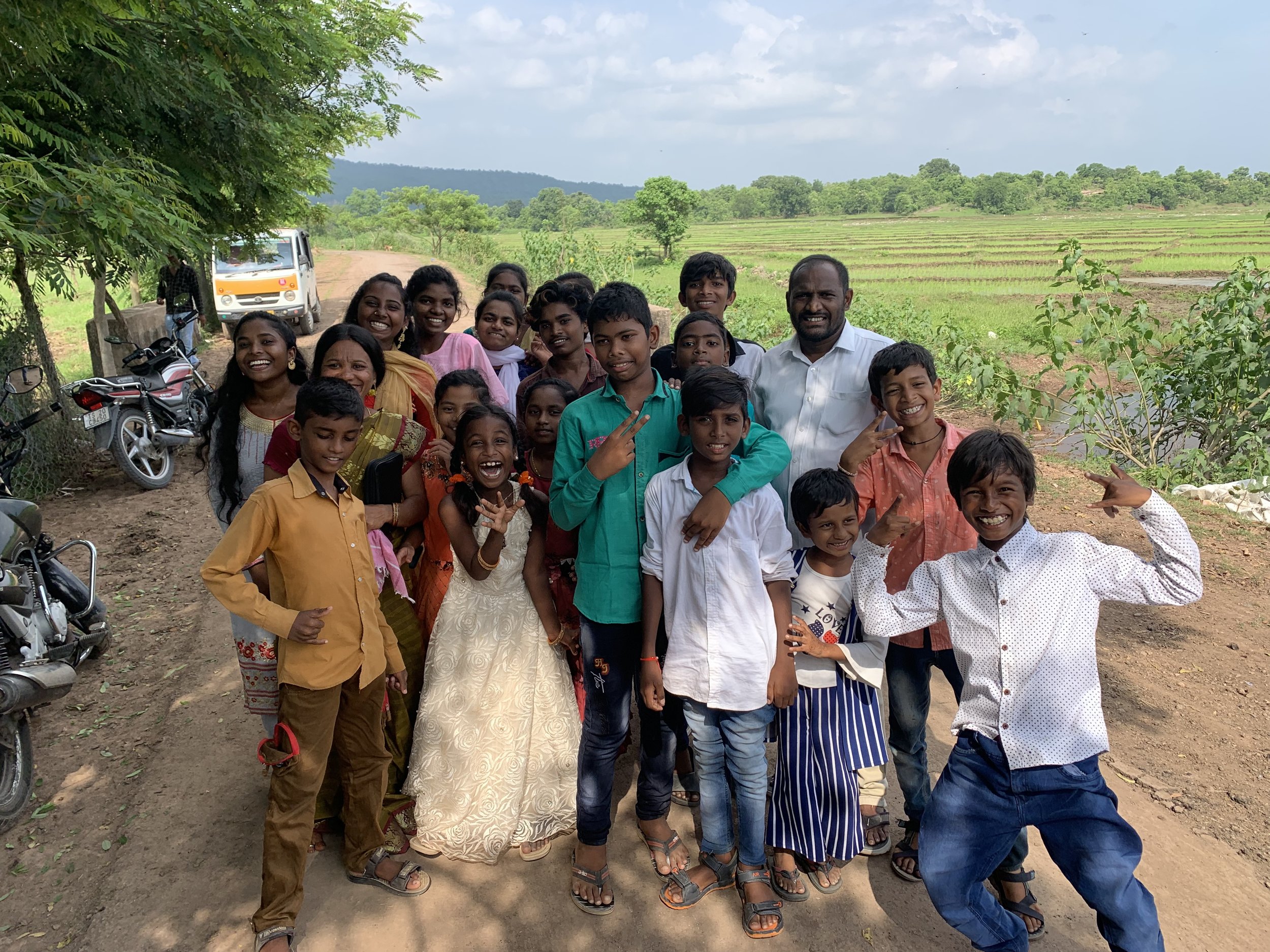 Group of smiling children and an adult standing on a rural dirt road with greenery and a vehicle in the background.