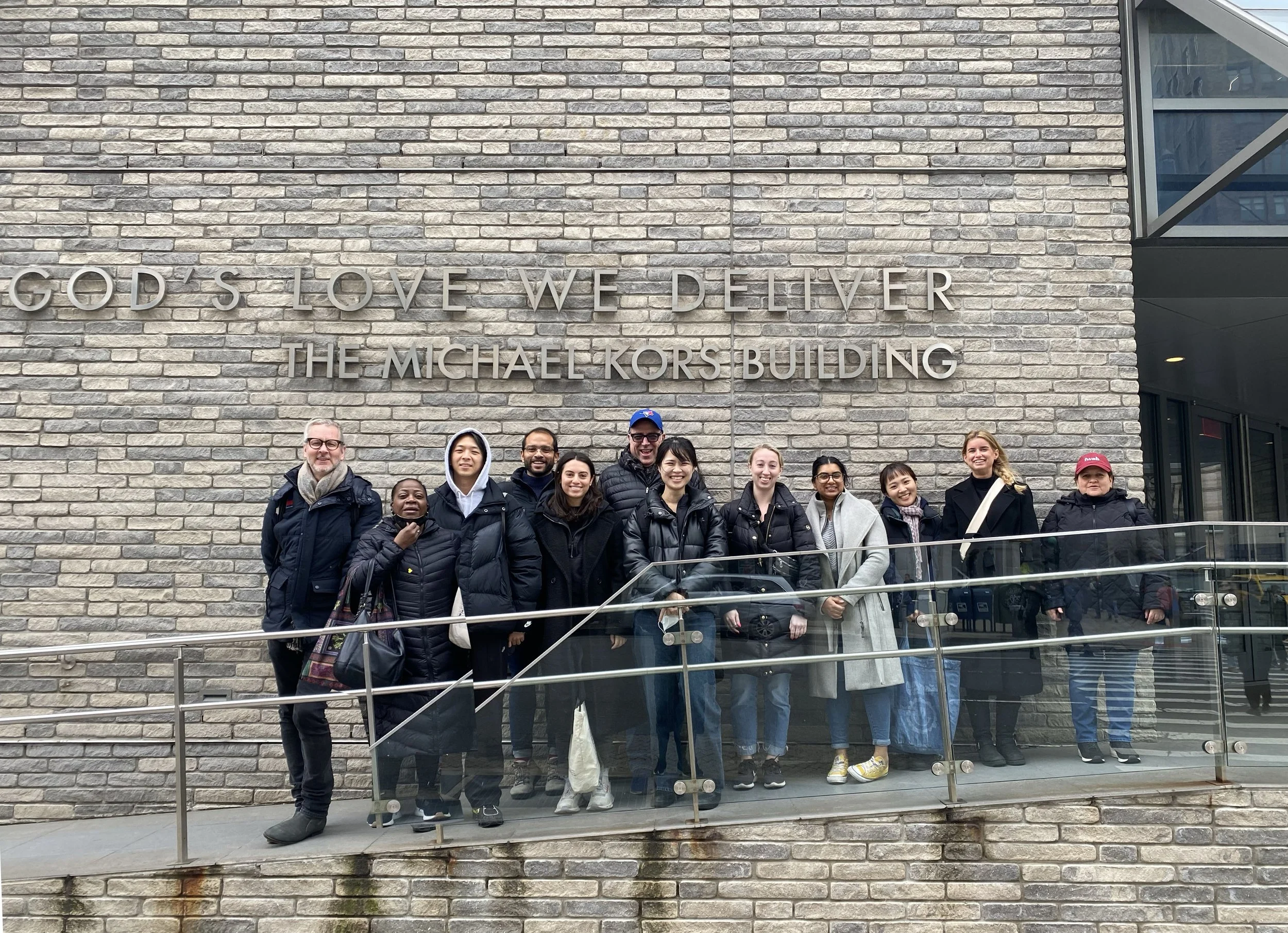 Group of diverse people posing for a photo outside a brick building with a sign that reads, 'GOD'S LOVE WE DELIVER THE MICHAEL KORS BUILDING.'