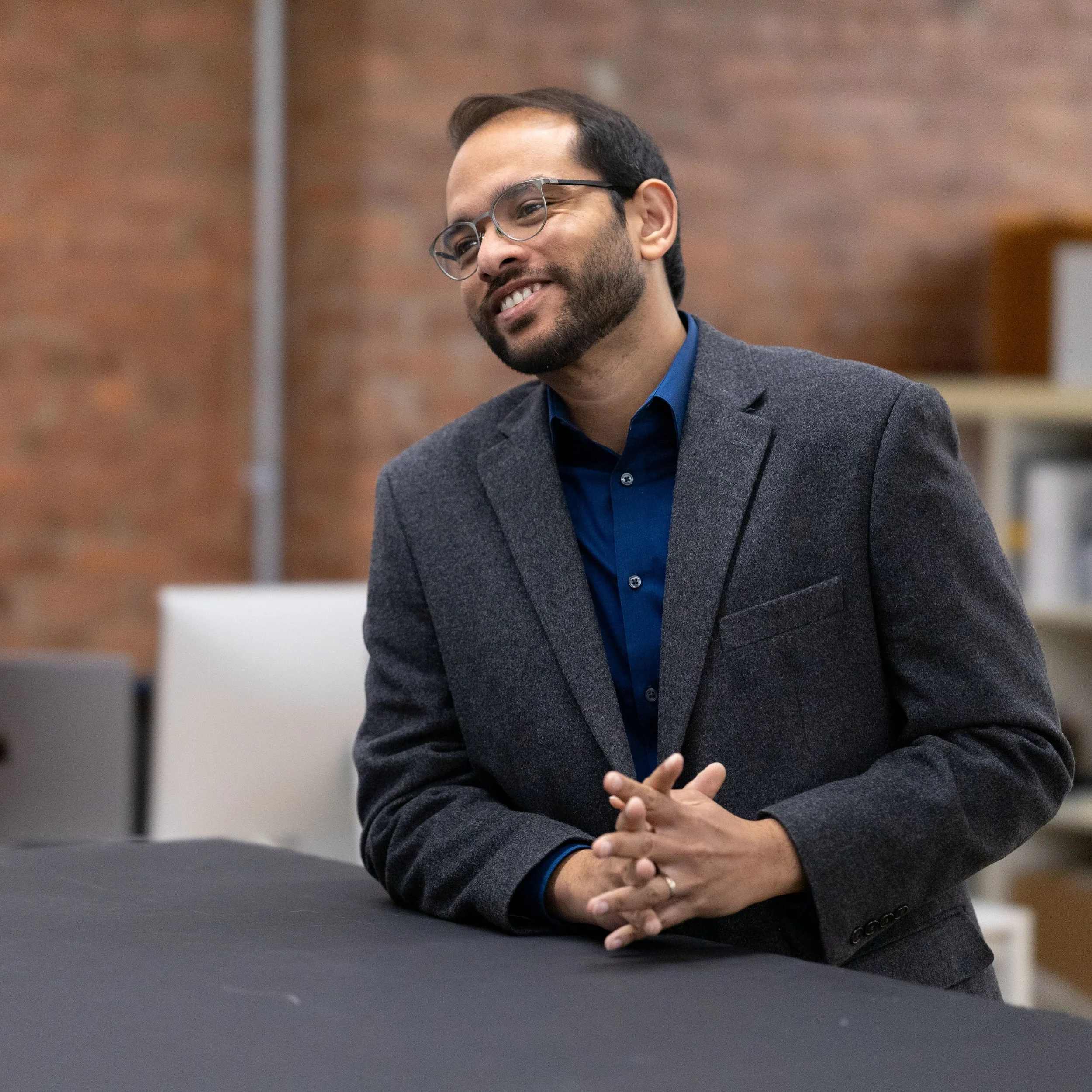 A man with glasses, a beard, and dark hair is smiling, wearing a dark gray suit and a blue shirt, with his hands clasped, standing in front of a brick wall and a white wall in an indoor setting.