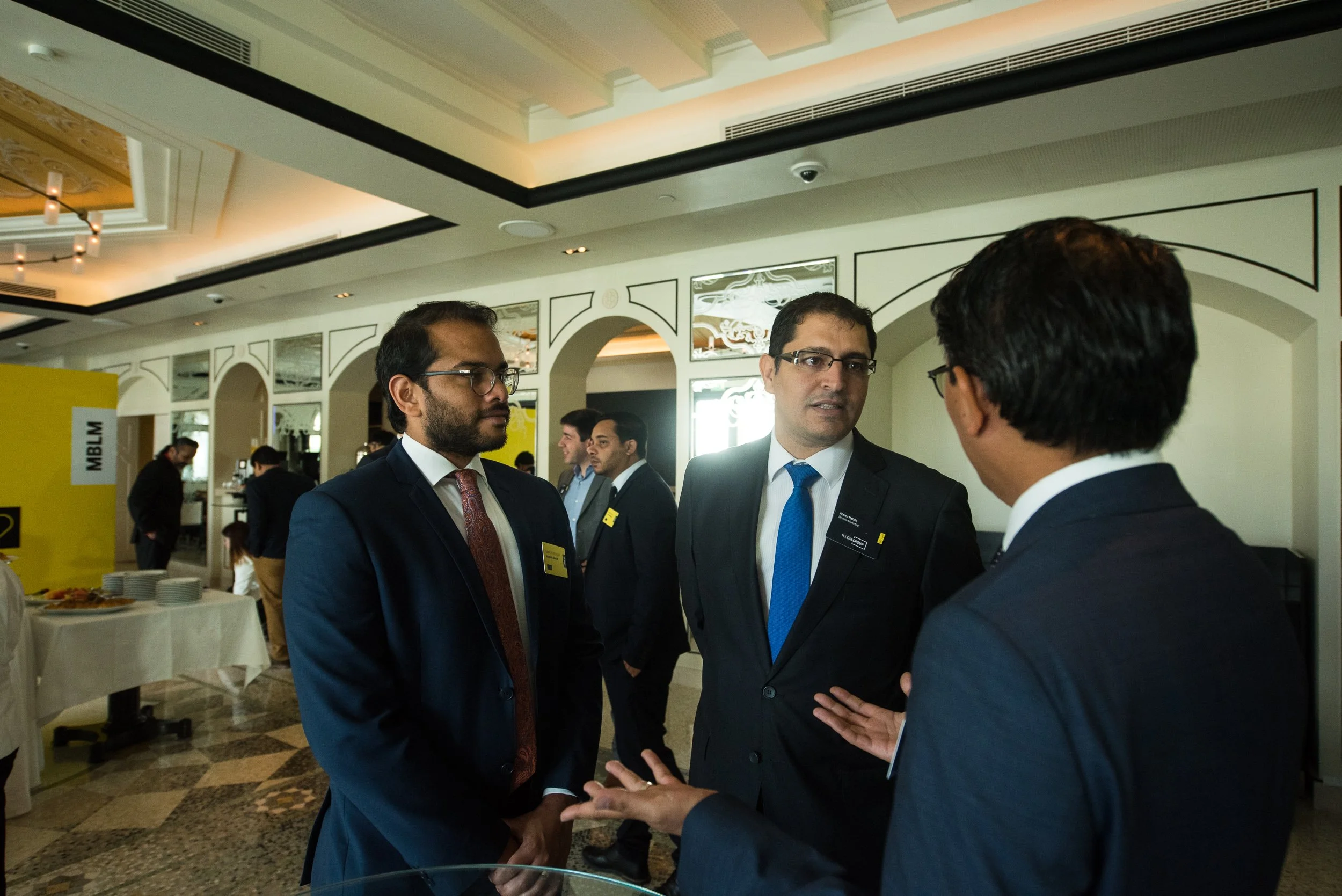 Four men in business suits engaged in conversation at a professional event. They are standing in a decorated room with white walls and a chandelier. There are other people and food stations in the background.
