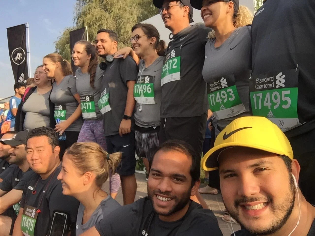 Group of people at a race event, some wearing race bibs with numbers, smiling and posing together for a photo. Some are standing, others sitting, with flags and trees in the background.