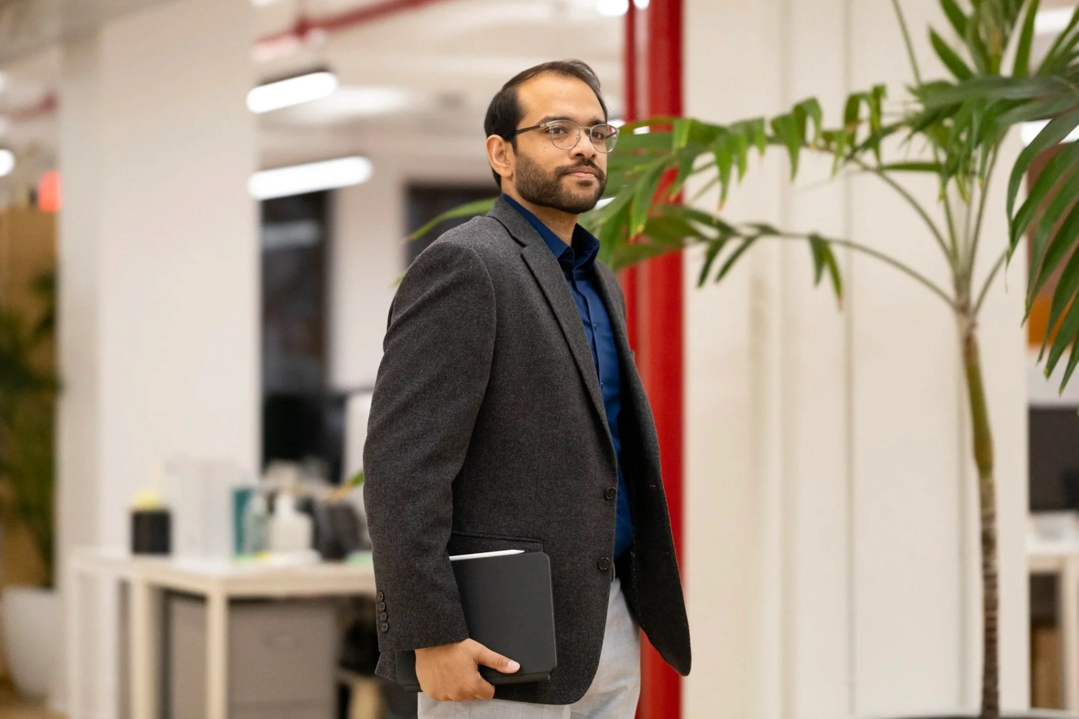 A man wearing glasses and a grey blazer standing in a modern office environment, holding a black laptop in his right hand.