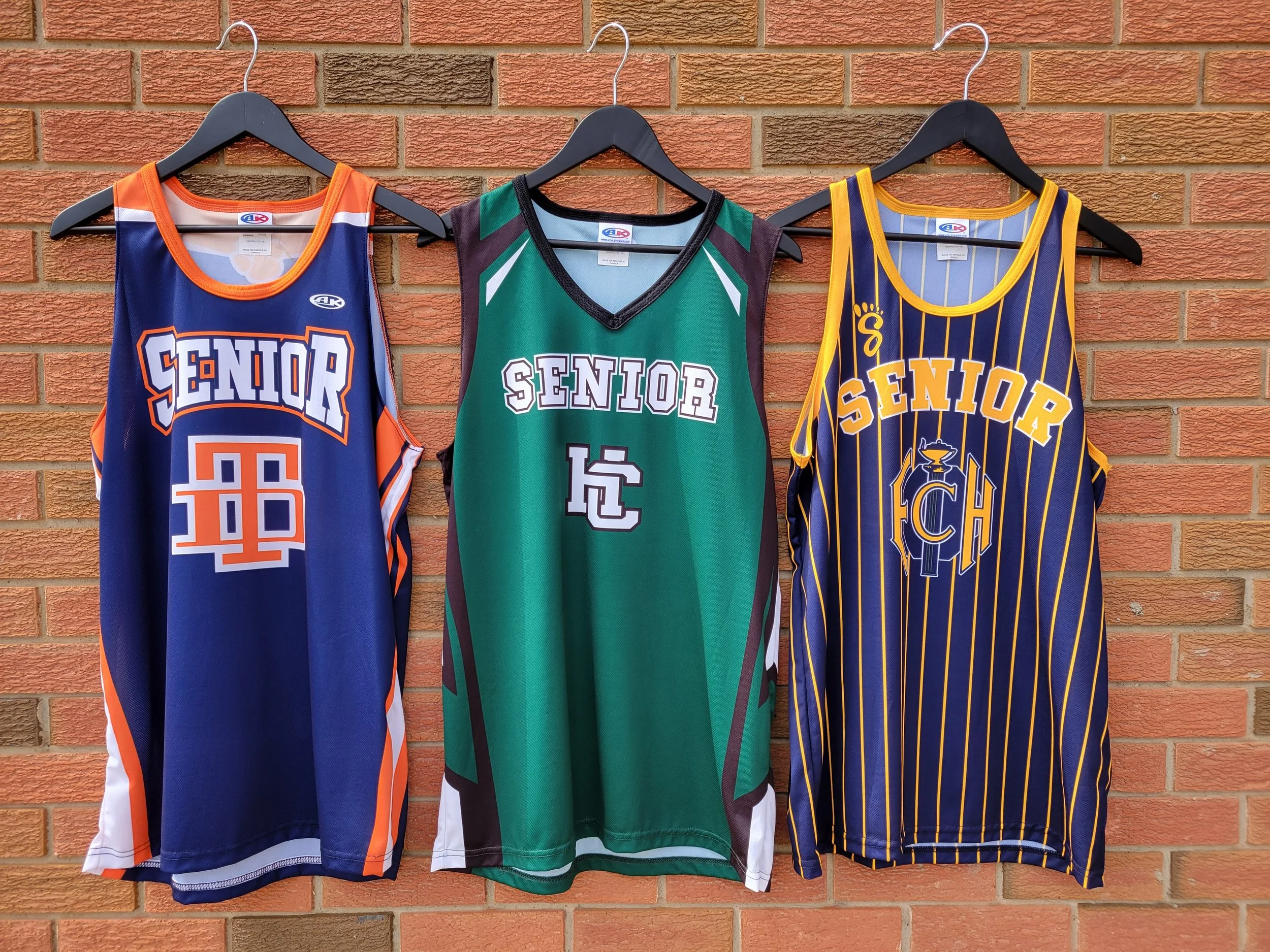 An image of three basketball jerseys hanging on a brick wall. First to the left is blue and orange, middle is green black and white, and the right jersey is yellow and blue with stripes.